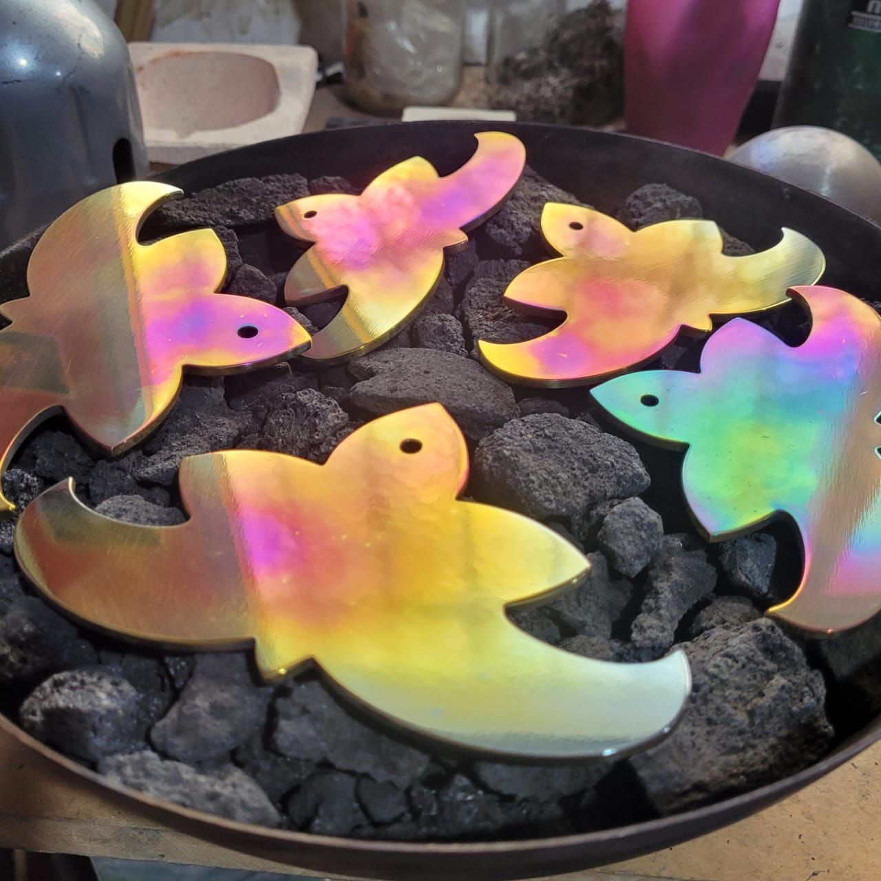 Seis recortes de metal iridiscentes con forma de pájaro en un recipiente de metal oscuro lleno de rocas negras.