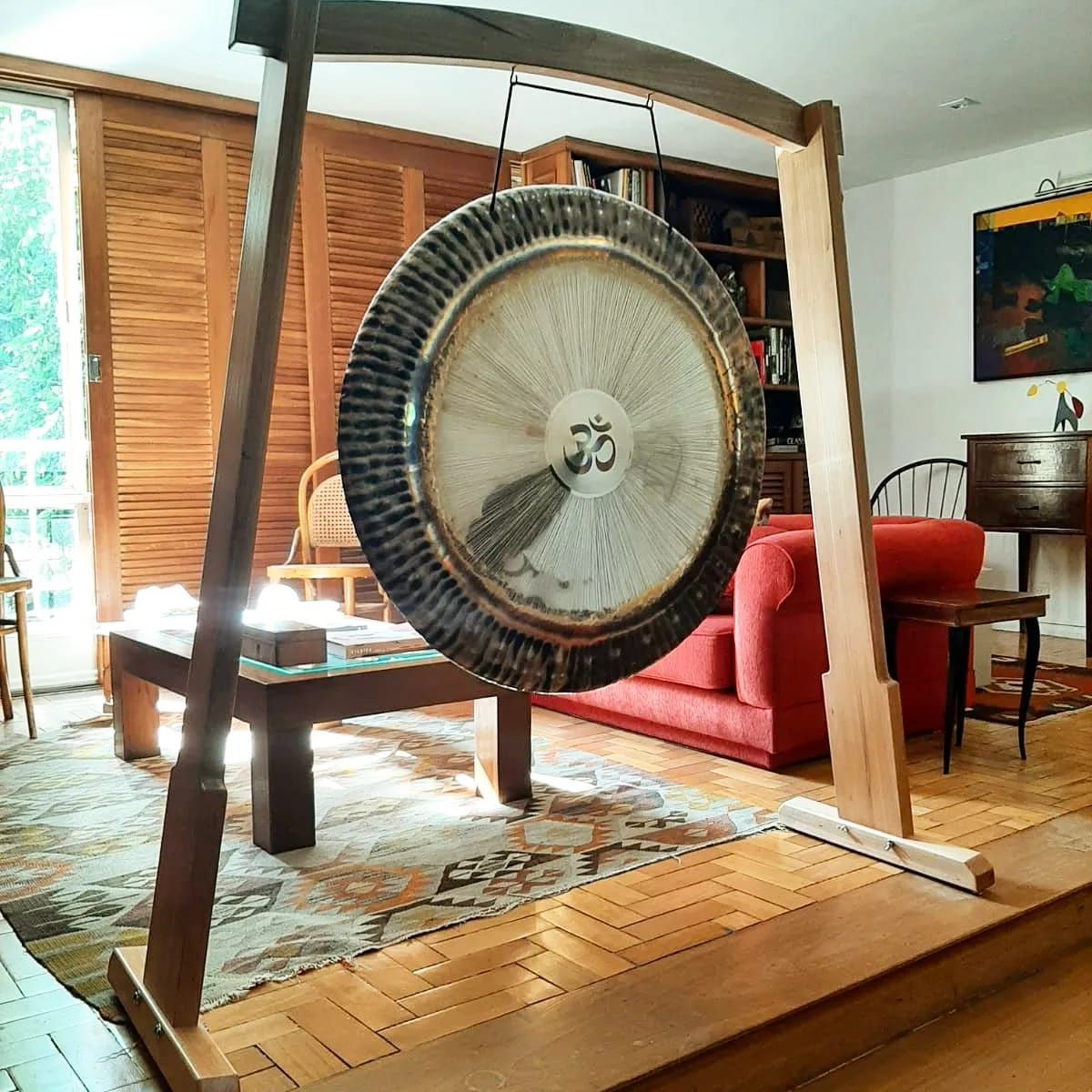 Un gran gong cuelga de un soporte de madera en una sala de estar. La luz del sol entra a raudales.