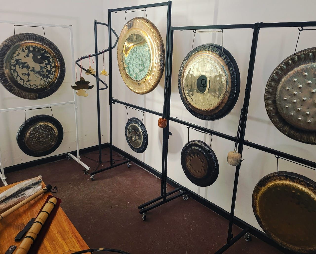 Gongs de varios tamaños colgados en soportes de metal blanco y negro en una habitación con una mesa de madera.