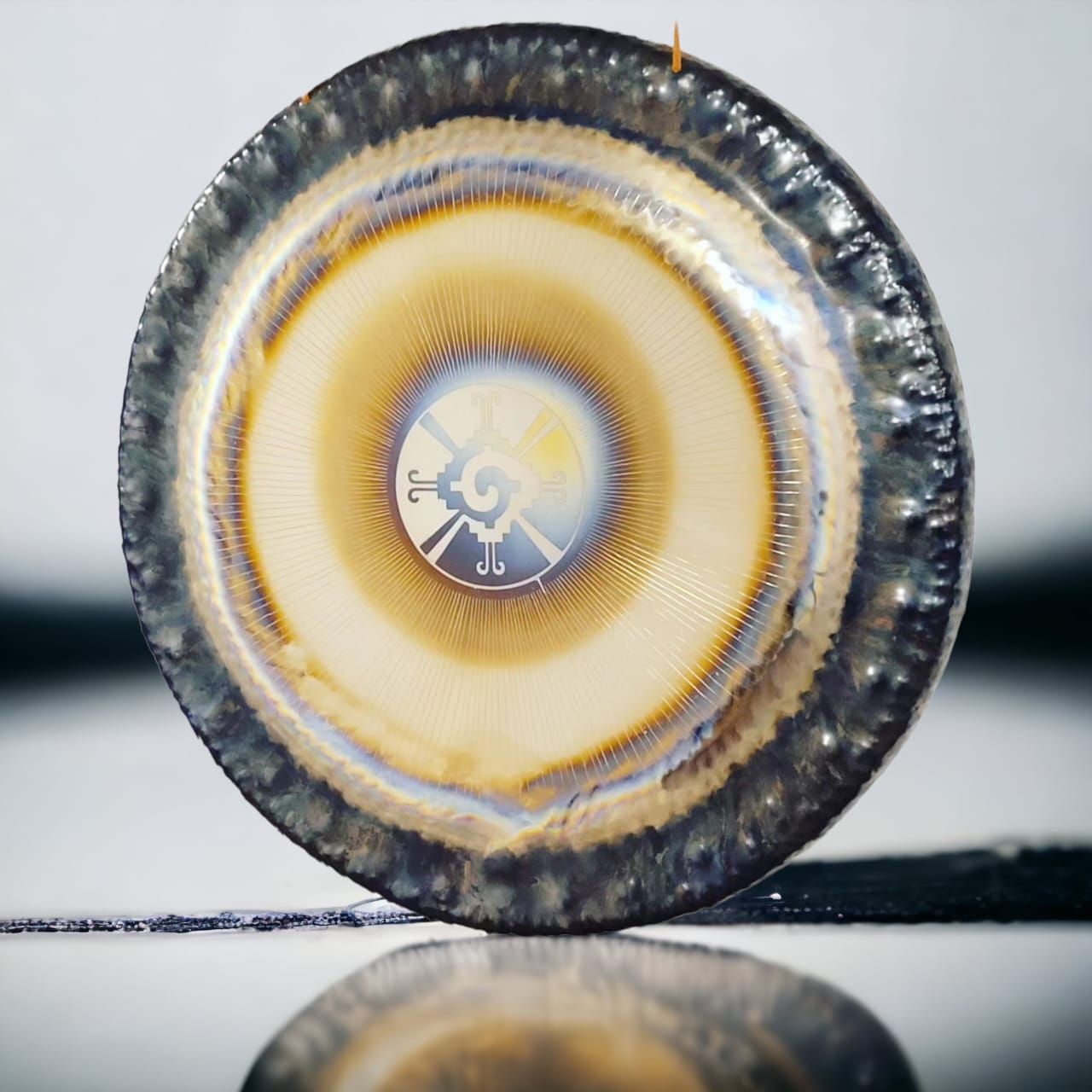 Gong circular de metal, con colores iridiscentes. Borde metálico y un pequeño diseño en el centro.