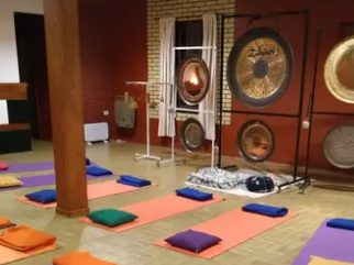 Estudio de yoga con gongs, esterillas y almohadas. Colores naranja, morado y azul. Paredes rojas y de ladrillo.