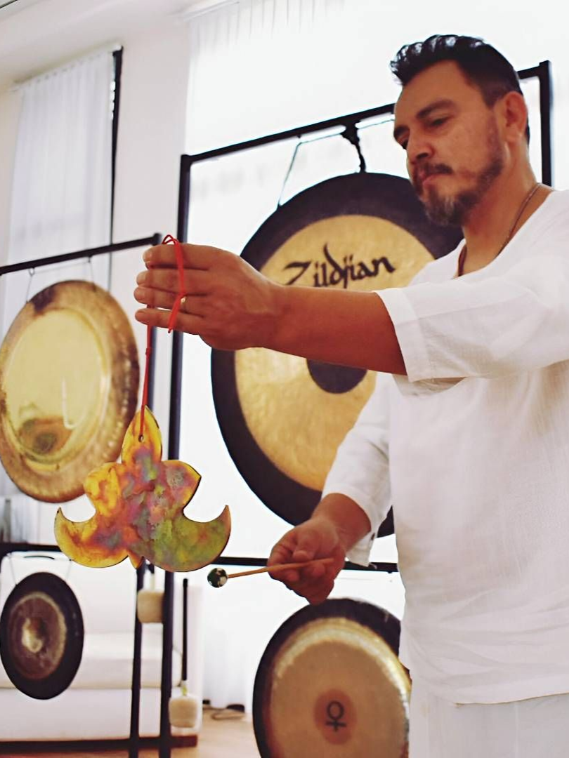 Hombre tocando gongs con un mazo en una habitación de paredes blancas con múltiples gongs.