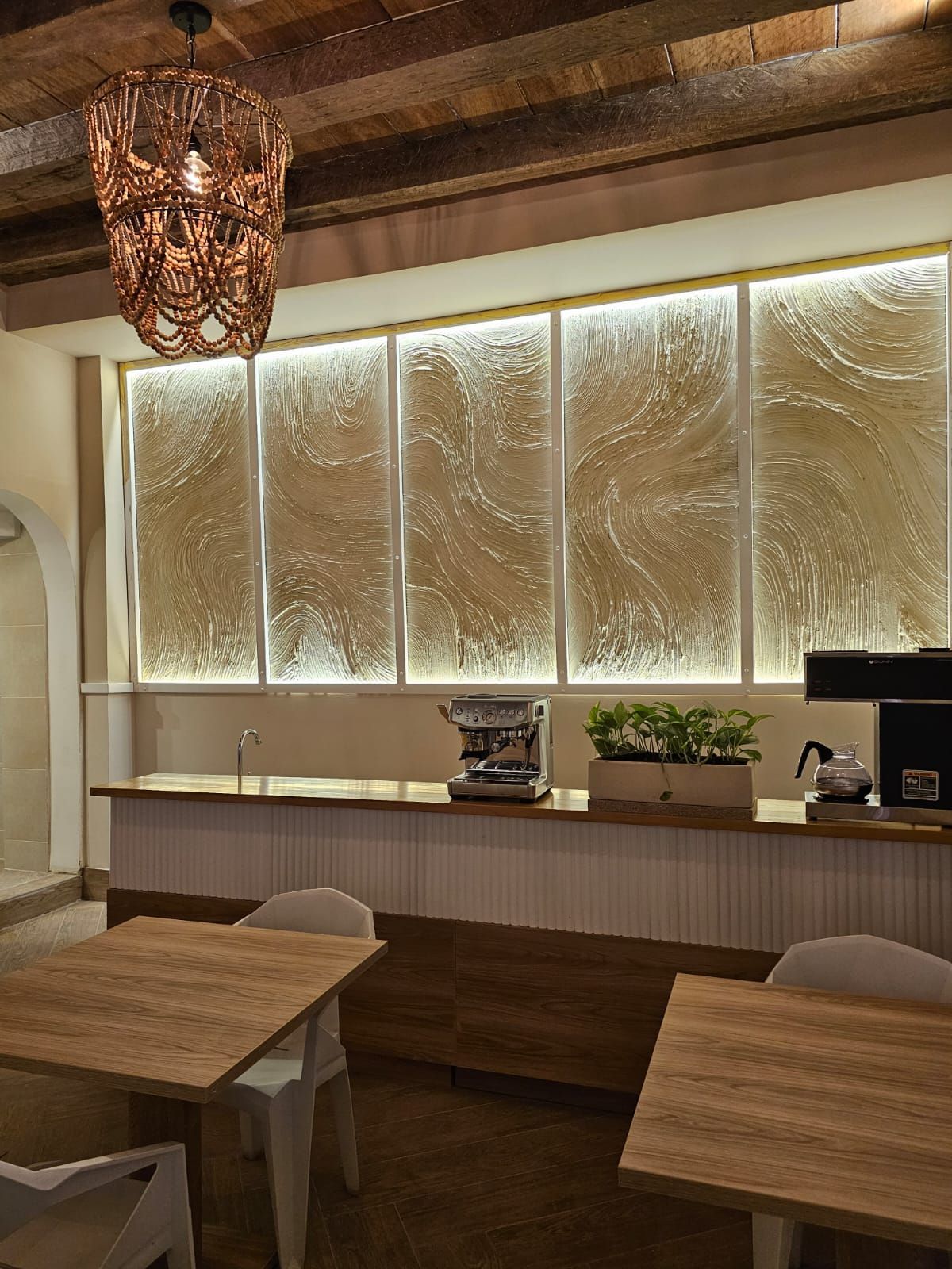 Interior of a cafe with a rustic vibe. Features wooden tables, a counter with coffee equipment, and a decorative back wall with integrated lighting.