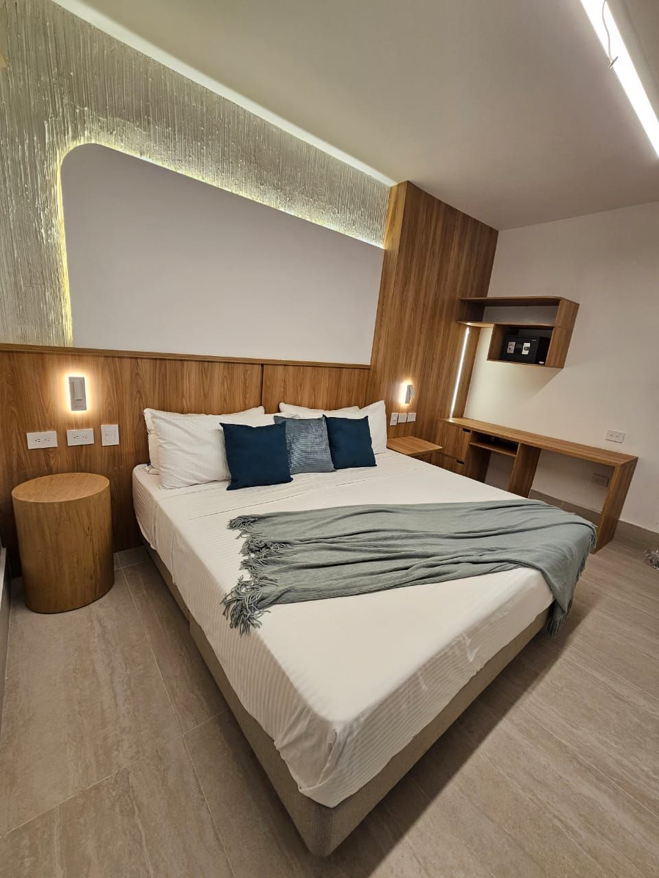 A modern hotel bedroom with a bed, wooden accents, and a desk. The bed has white linens, blue pillows, and a blue throw.