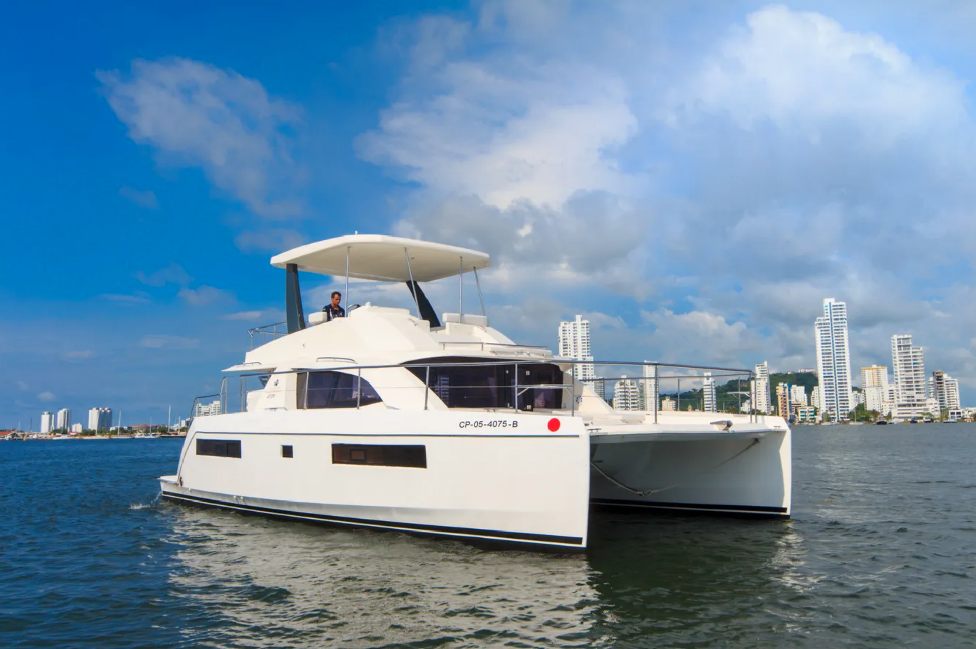 White catamaran sailing on water with a cityscape in the background under a blue sky.