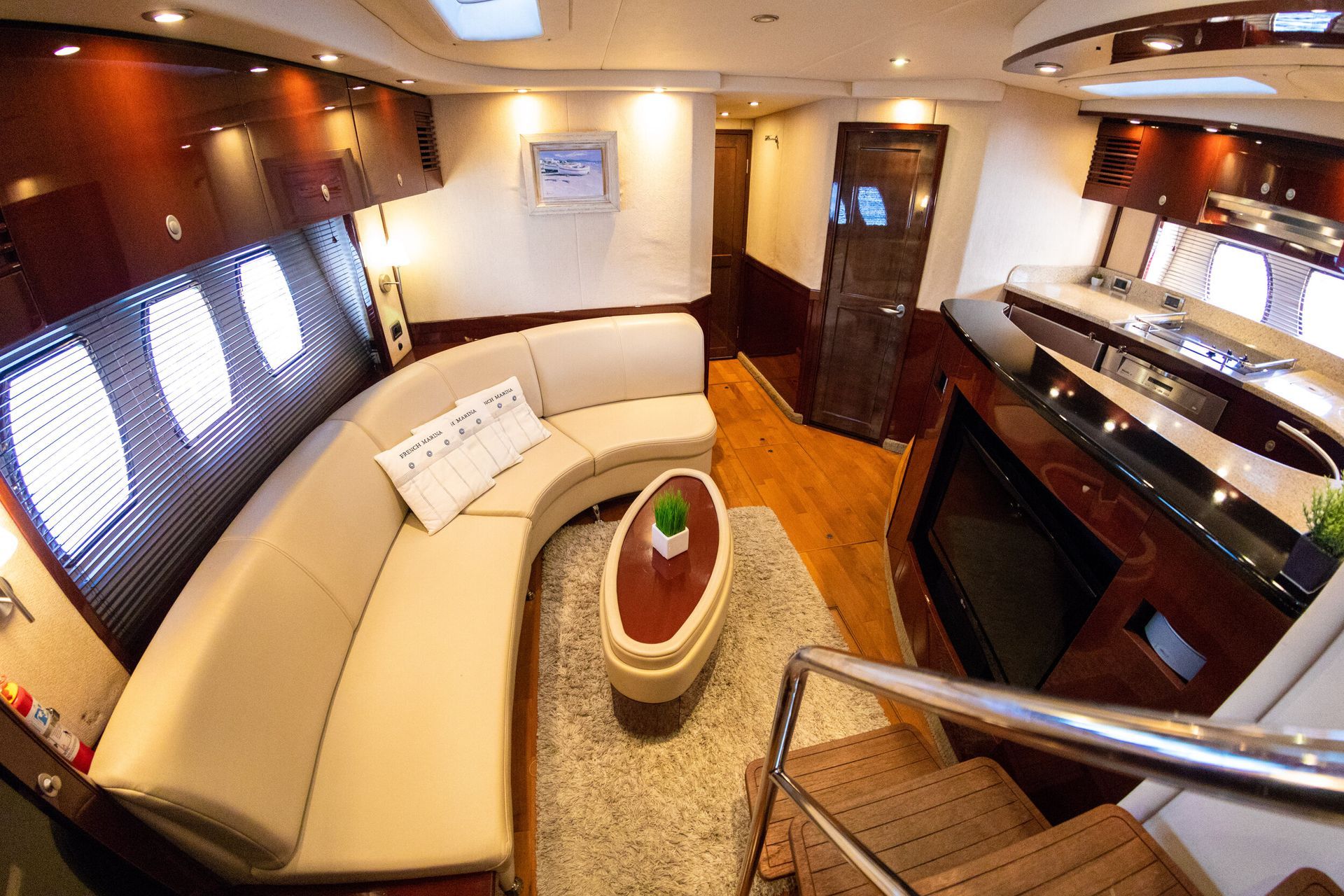 Interior of a yacht with a curved beige sofa, small oval coffee table, and staircase leading to a lower level. Dark wood paneling and a fireplace add to the luxurious setting.