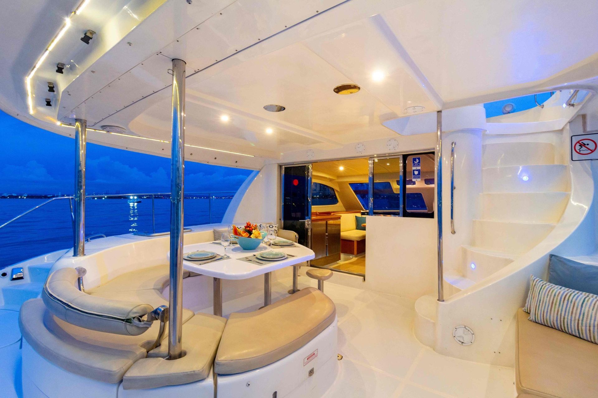 Boat interior with curved seating around a table, a spiral staircase, and a view into the boat's interior at night.