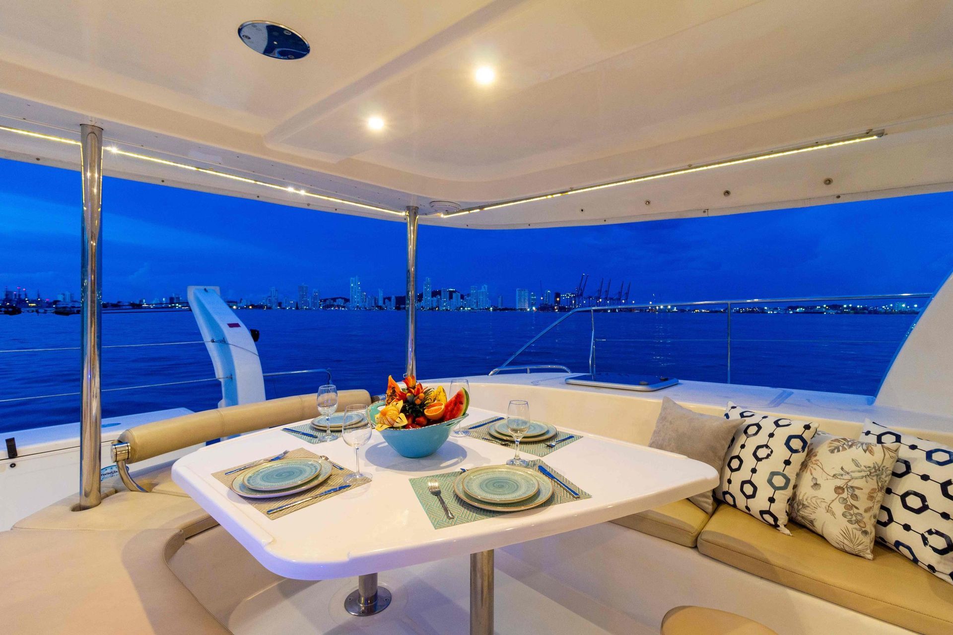 Outdoor dining area on a yacht at dusk, with a table set for a meal, and city skyline in the background.