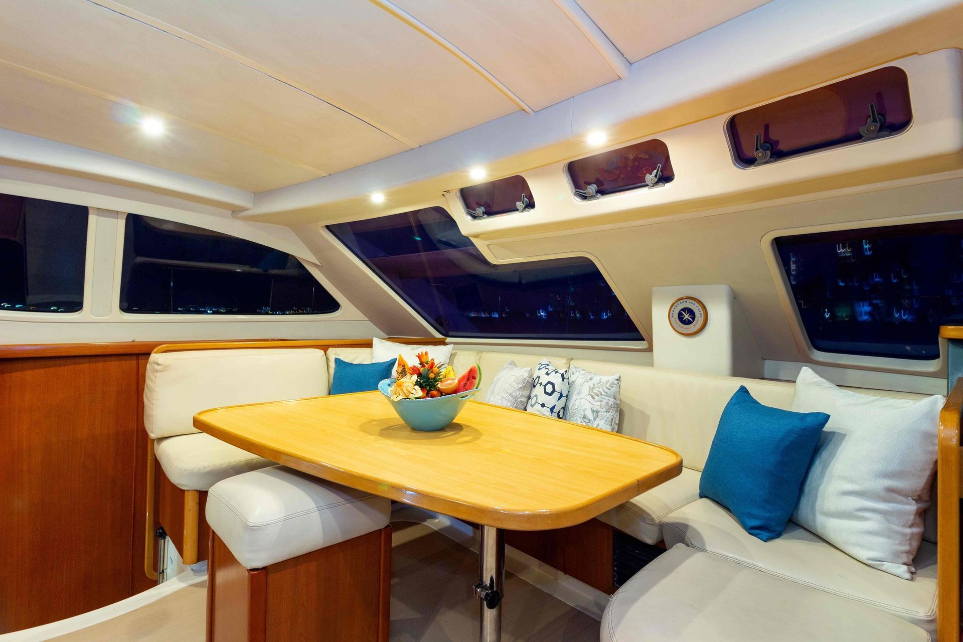 Interior of a sailboat with a wooden table, built-in seating, and windows. Turquoise and white pillows and a flower bowl add color.