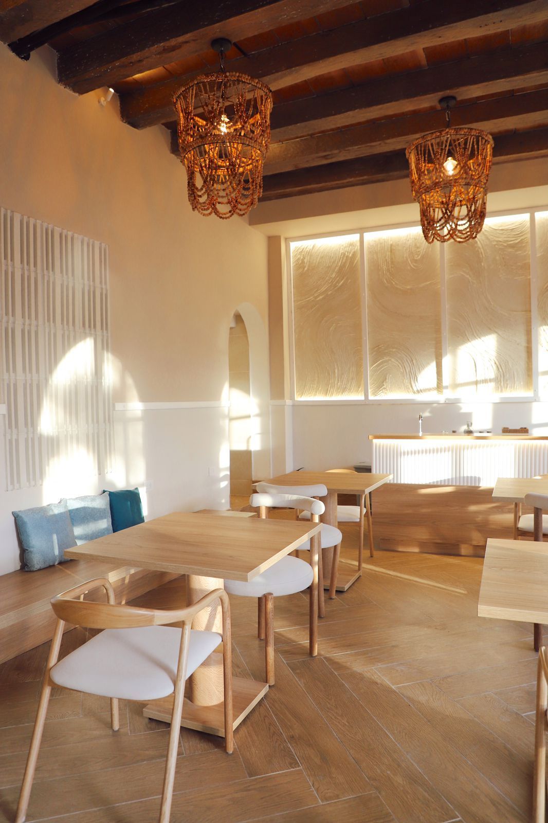 A light-filled restaurant interior featuring wooden tables and chairs, woven pendant lights, and a built-in bench with blue cushions.