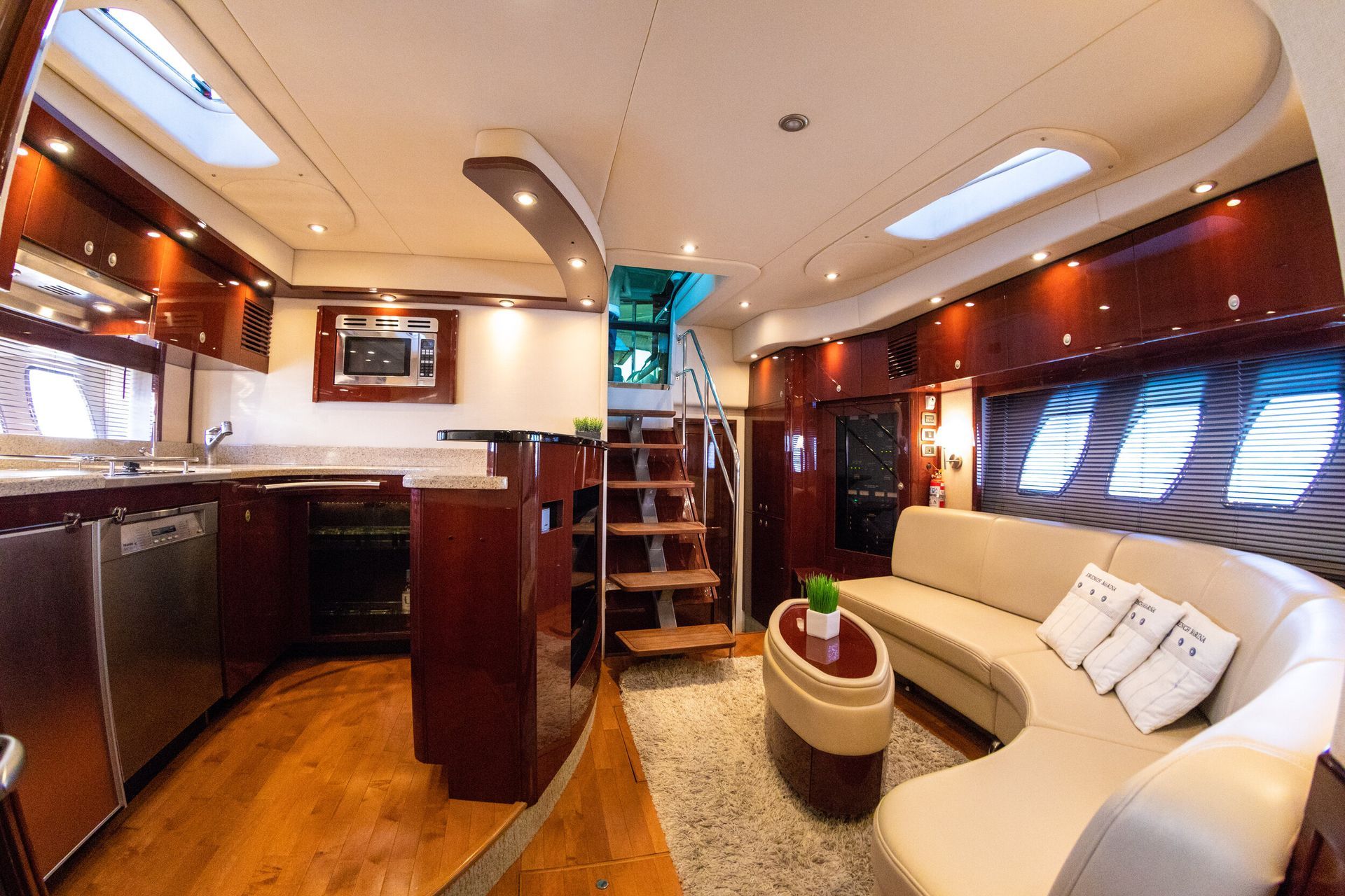 Interior of a yacht with a kitchen on the left, a stairway, and a curved cream-colored sofa. Wooden floors and mahogany cabinets.