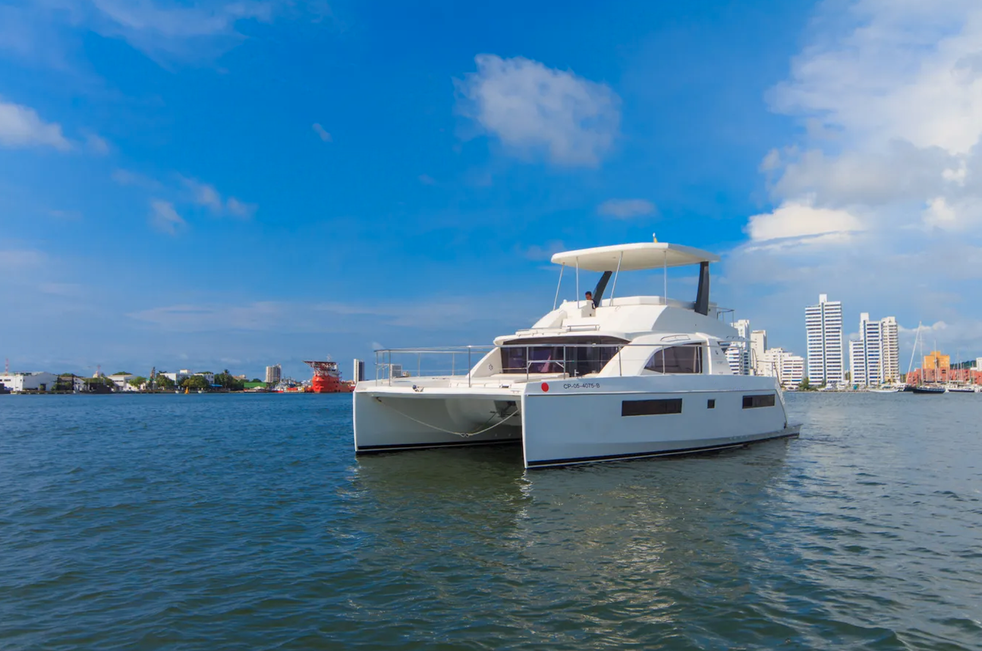 White catamaran sailing on blue water under a partly cloudy sky, city skyline in the background.