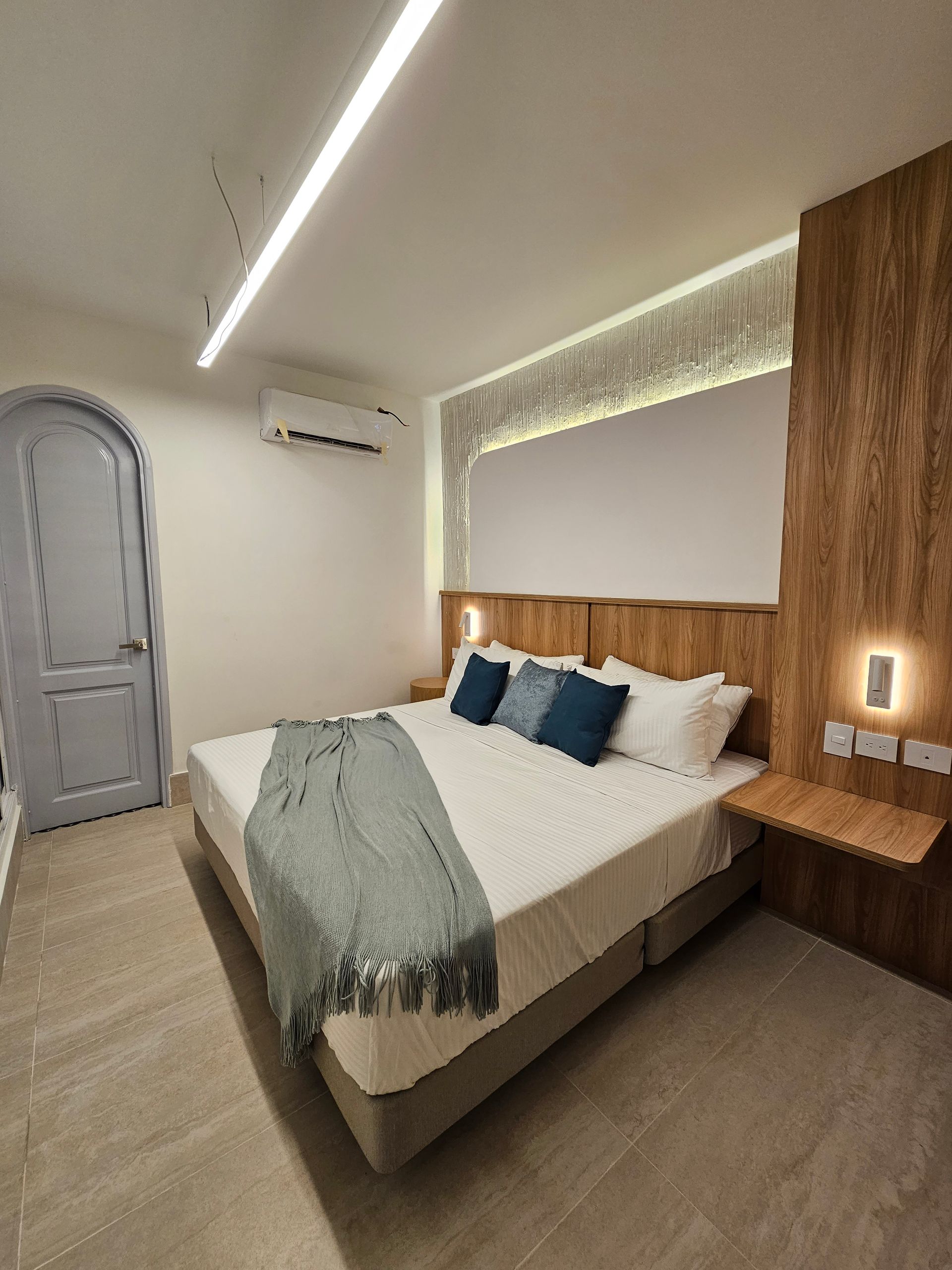 A modern hotel bedroom with a king-sized bed, wooden headboard, and an arched blue door.