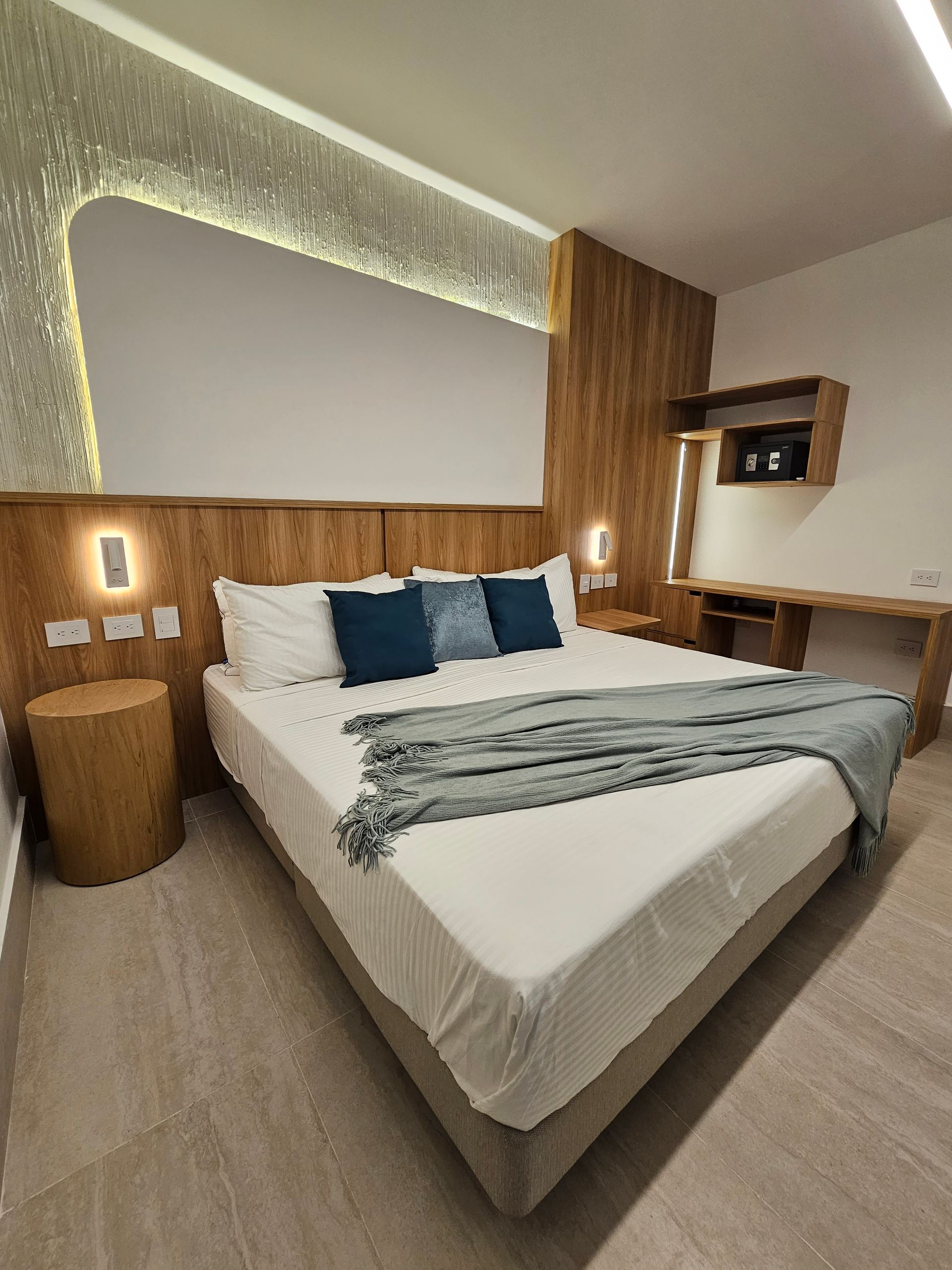 Modern hotel bedroom with a bed, wooden accents, and a desk. The room has light-colored walls and floor.