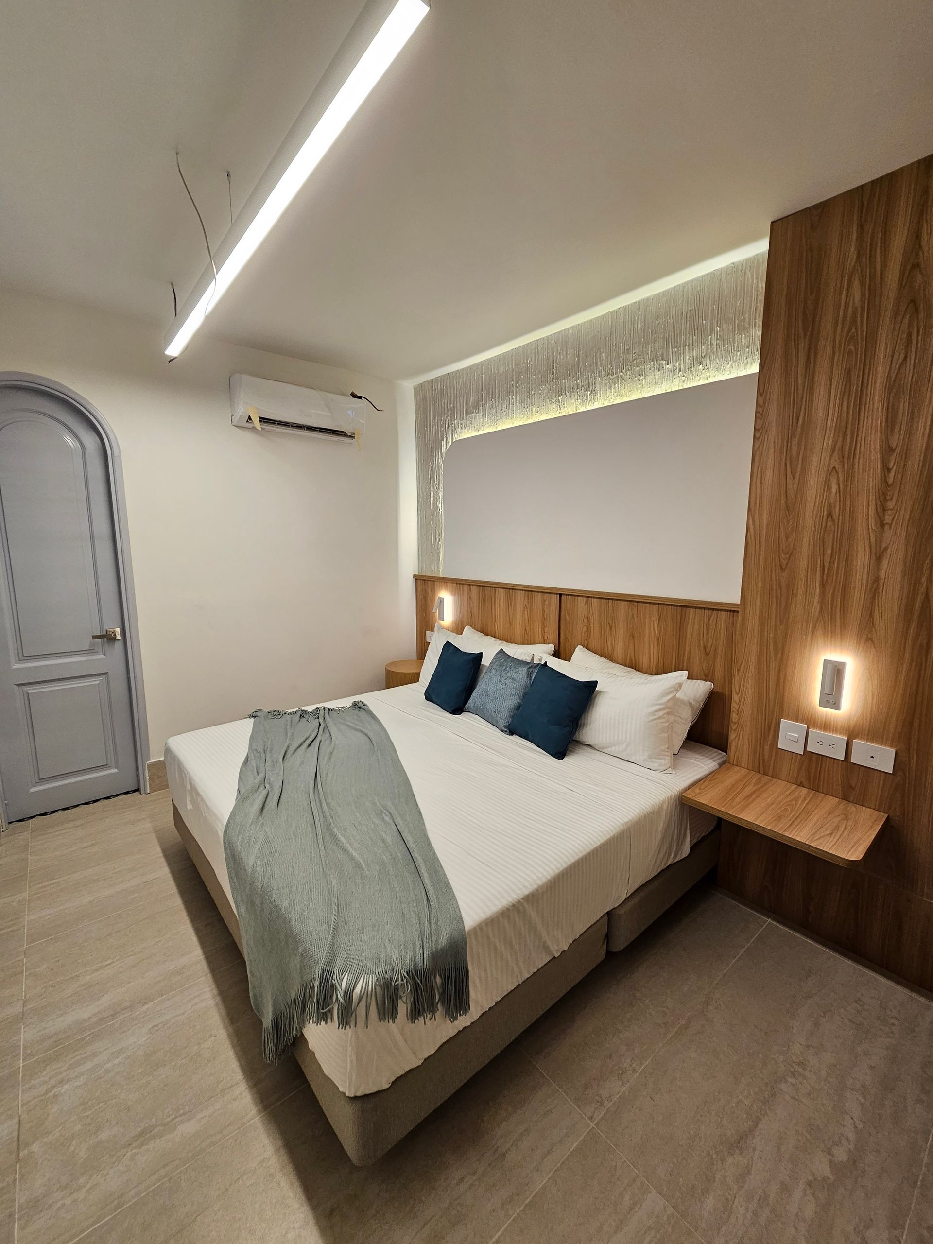 Modern bedroom with a large bed, wooden accents, and a blue blanket. Door is on the left, a small nightstand on the right.