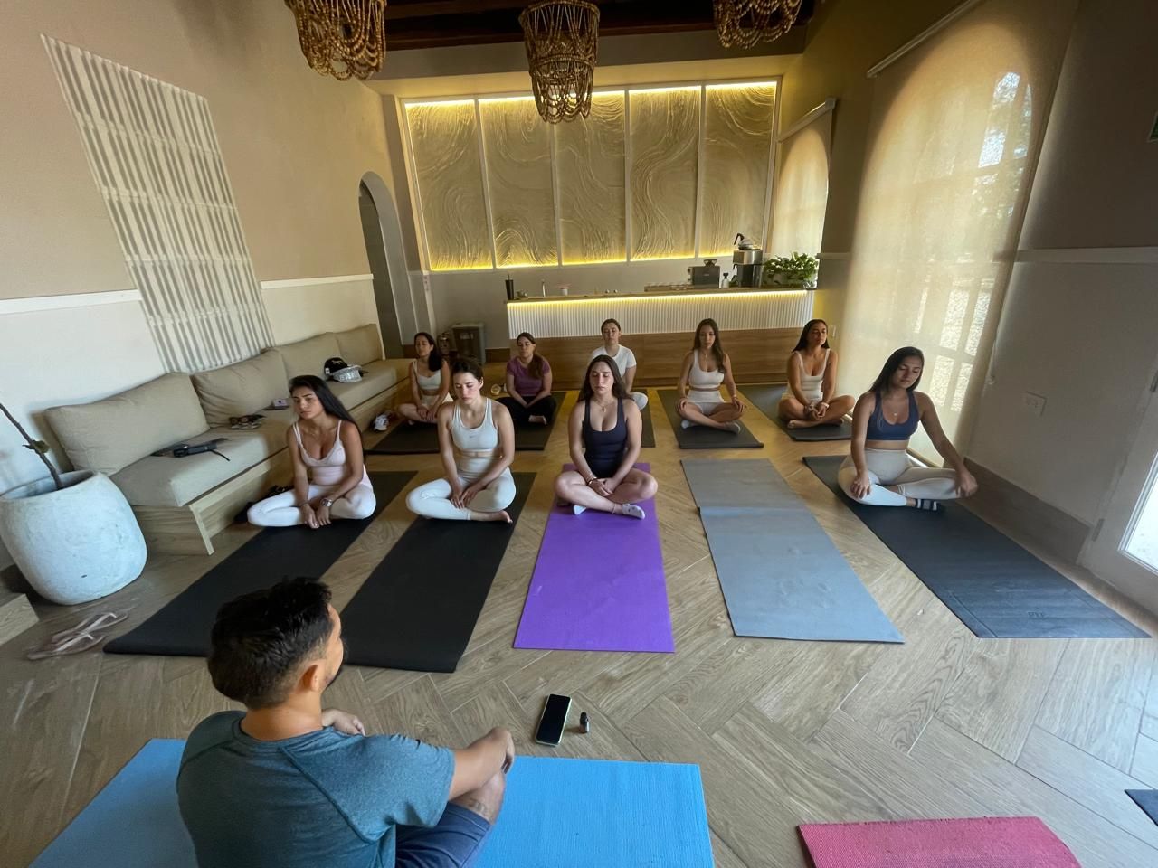 A group of people are sitting on yoga mats, meditating in a bright room. A person is facing the group, possibly instructing.