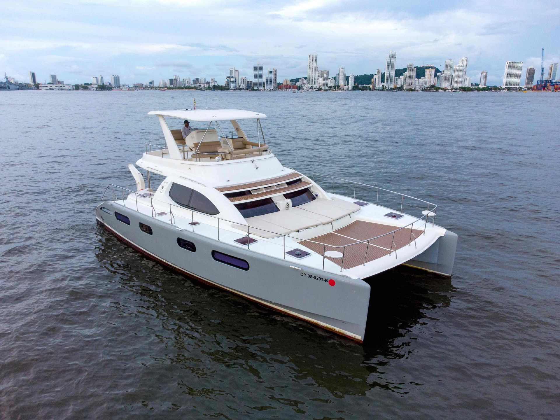 A white catamaran boat sails on a body of water with a city skyline visible in the background.