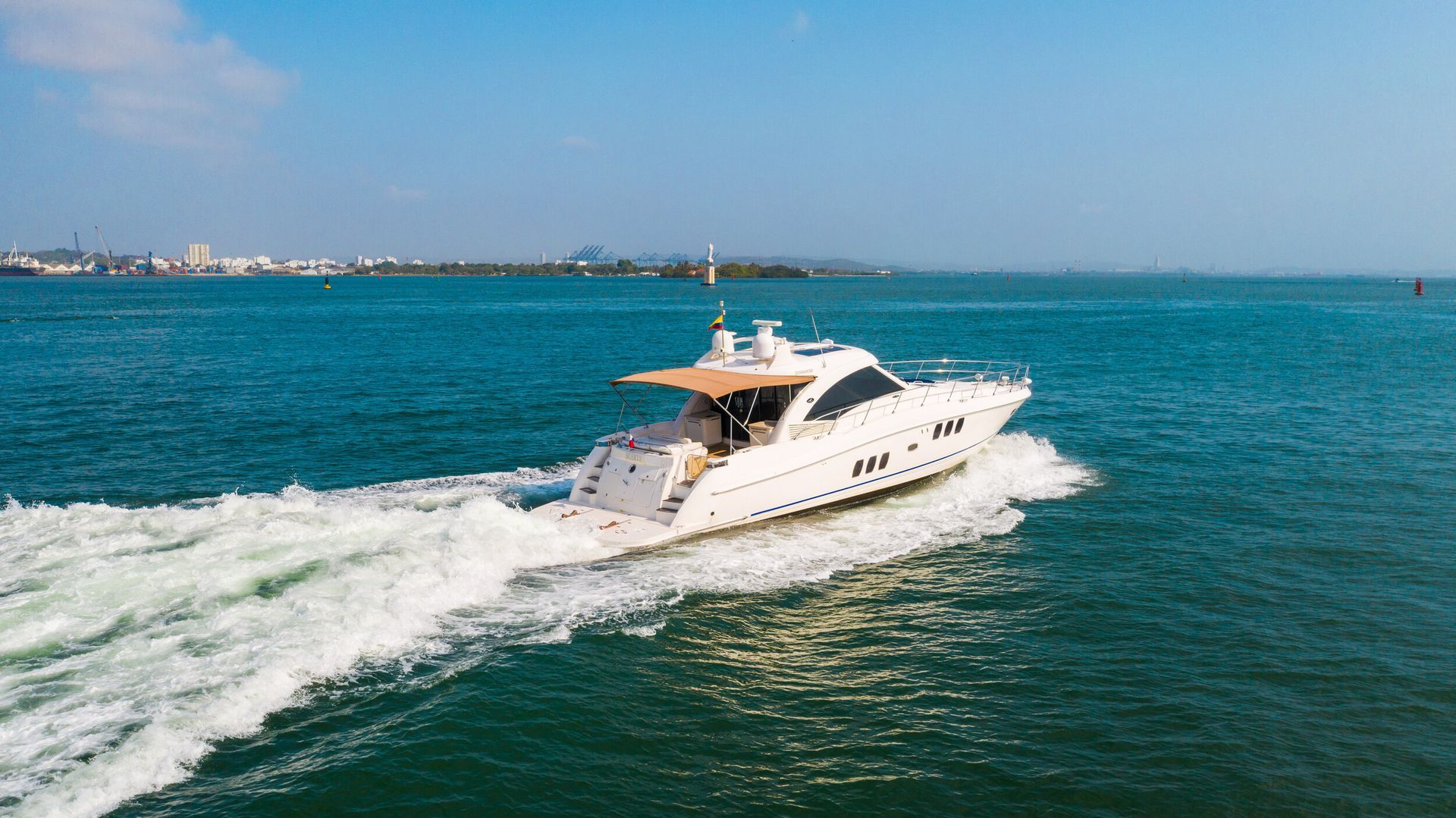 White yacht sailing on blue water, leaving a white wake. In the background, a city skyline is visible.
