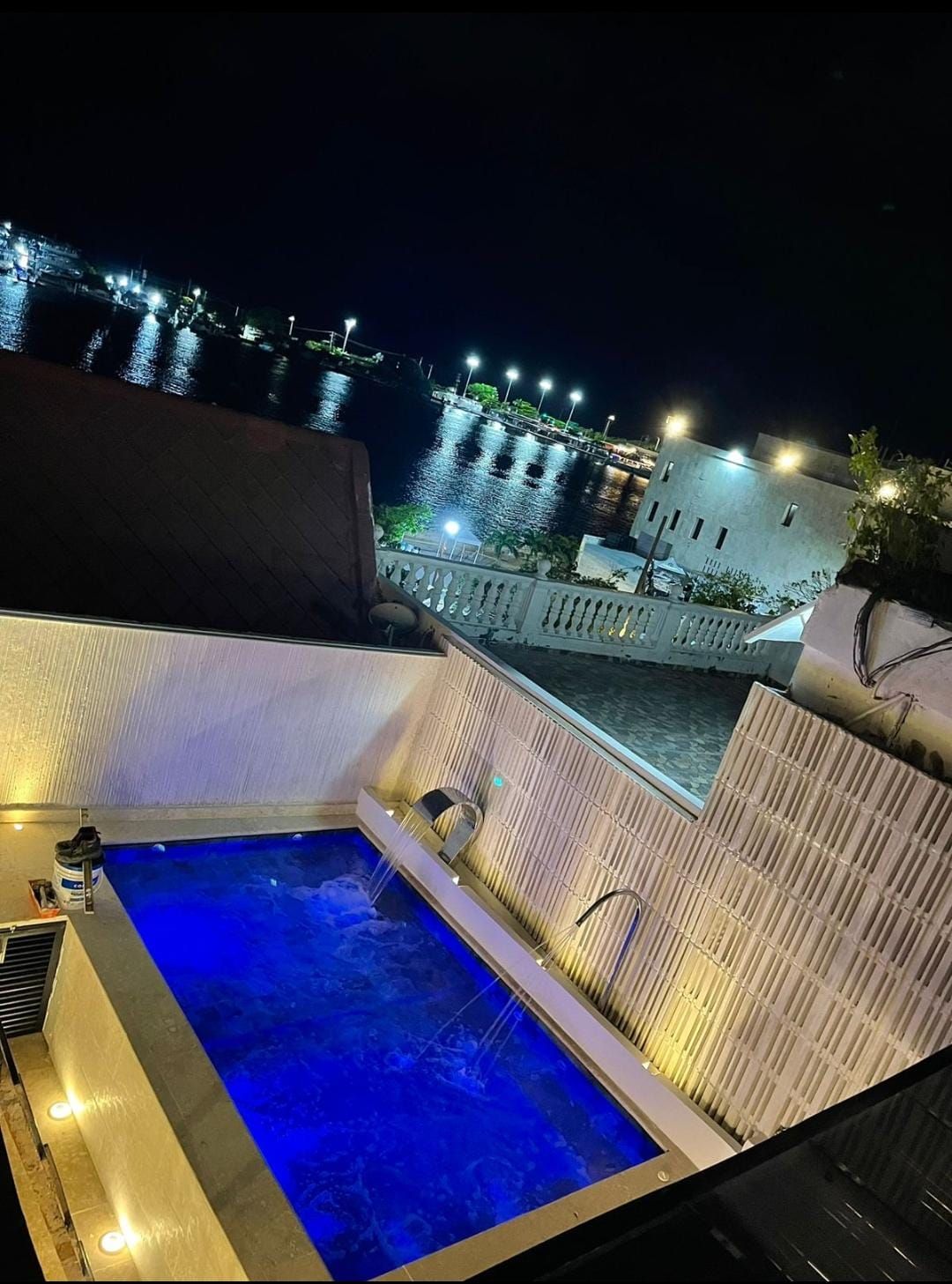Night view of a pool with illuminated blue water, overlooking a waterfront with lights reflecting in the dark water.
