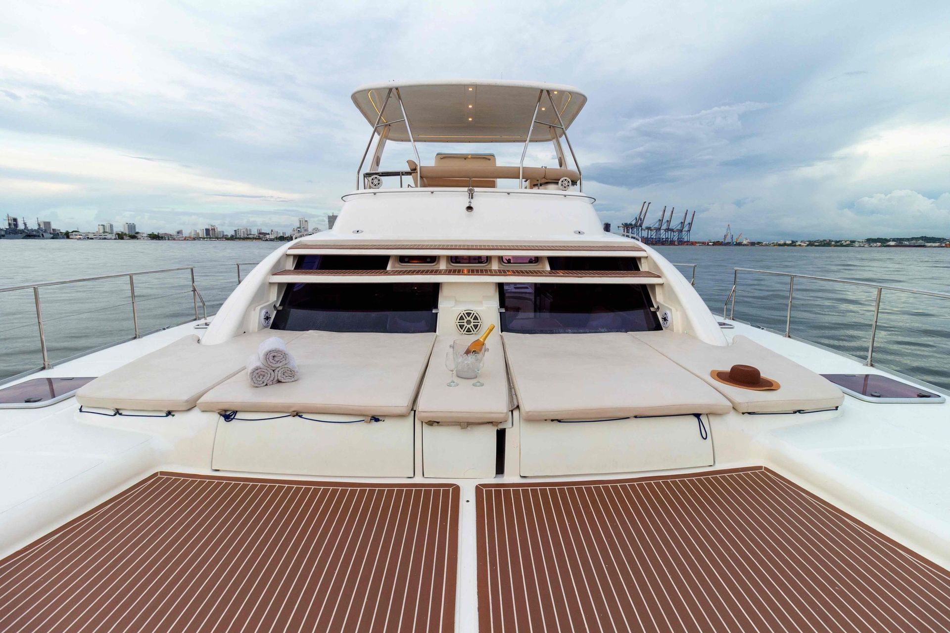 A white catamaran with cushioned sunbeds, a shaded seating area, and a view of the water and a distant cityscape.