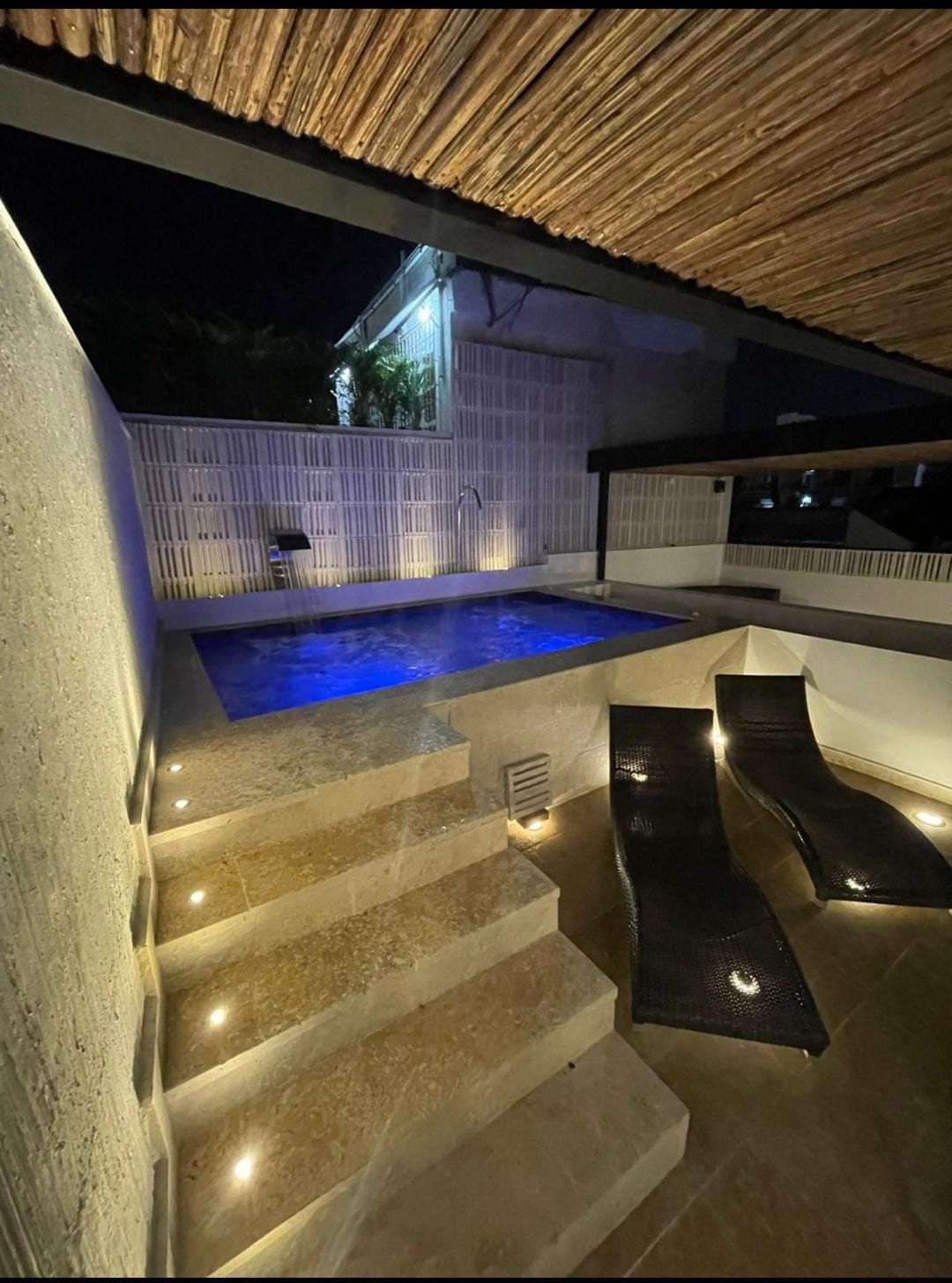 A small, illuminated pool at night with two lounge chairs on a terrace. Stone steps lead down to the pool area.