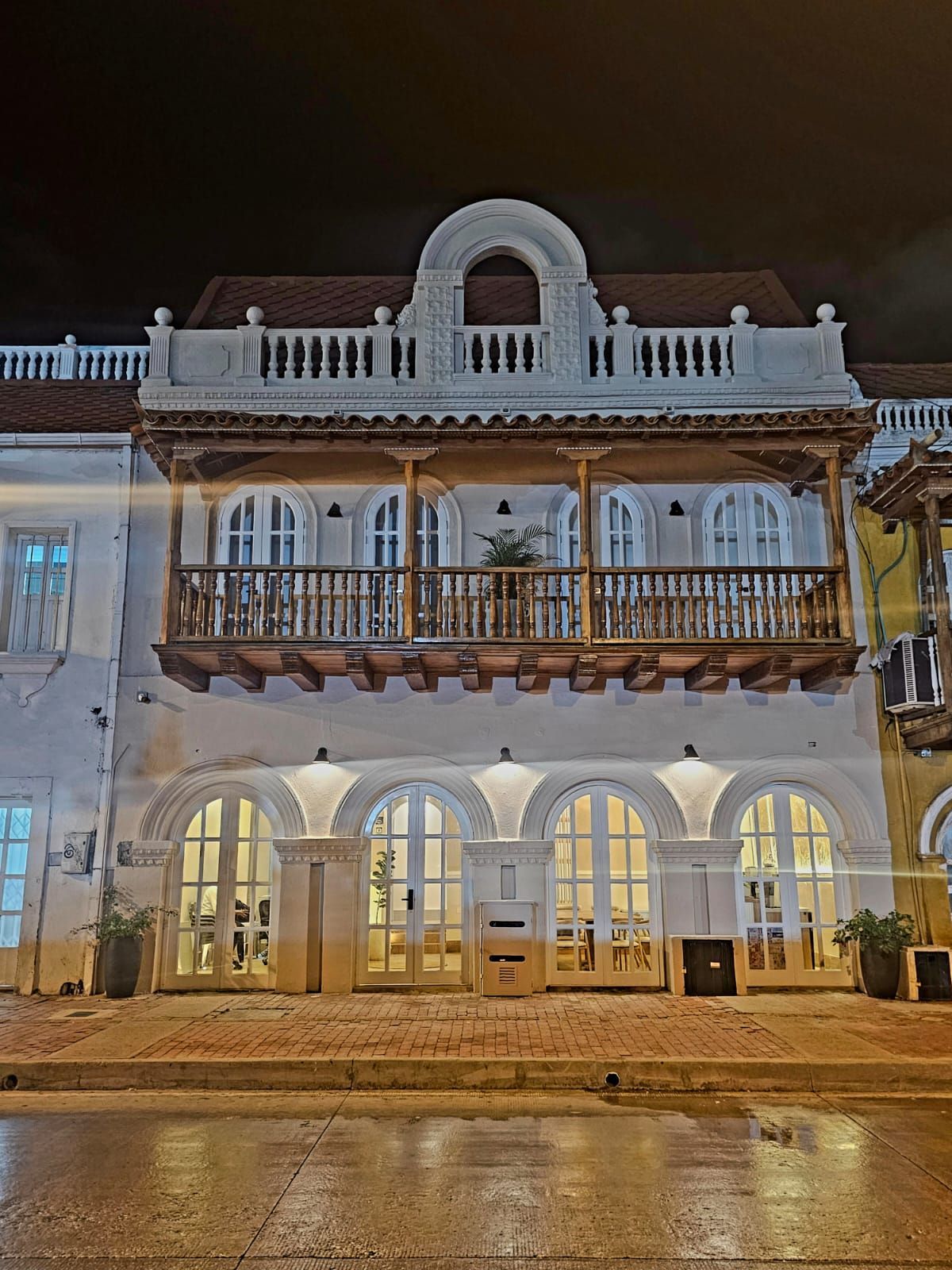 Night shot of a white colonial building in Cartagena, Colombia, featuring arched windows and a wooden balcony with intricate detailing.