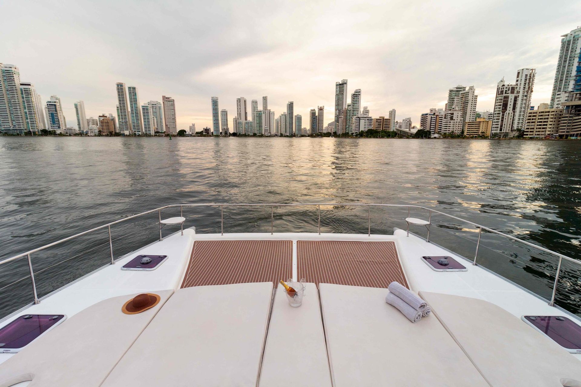 A catamaran deck with lounge seating faces a city skyline at dusk.