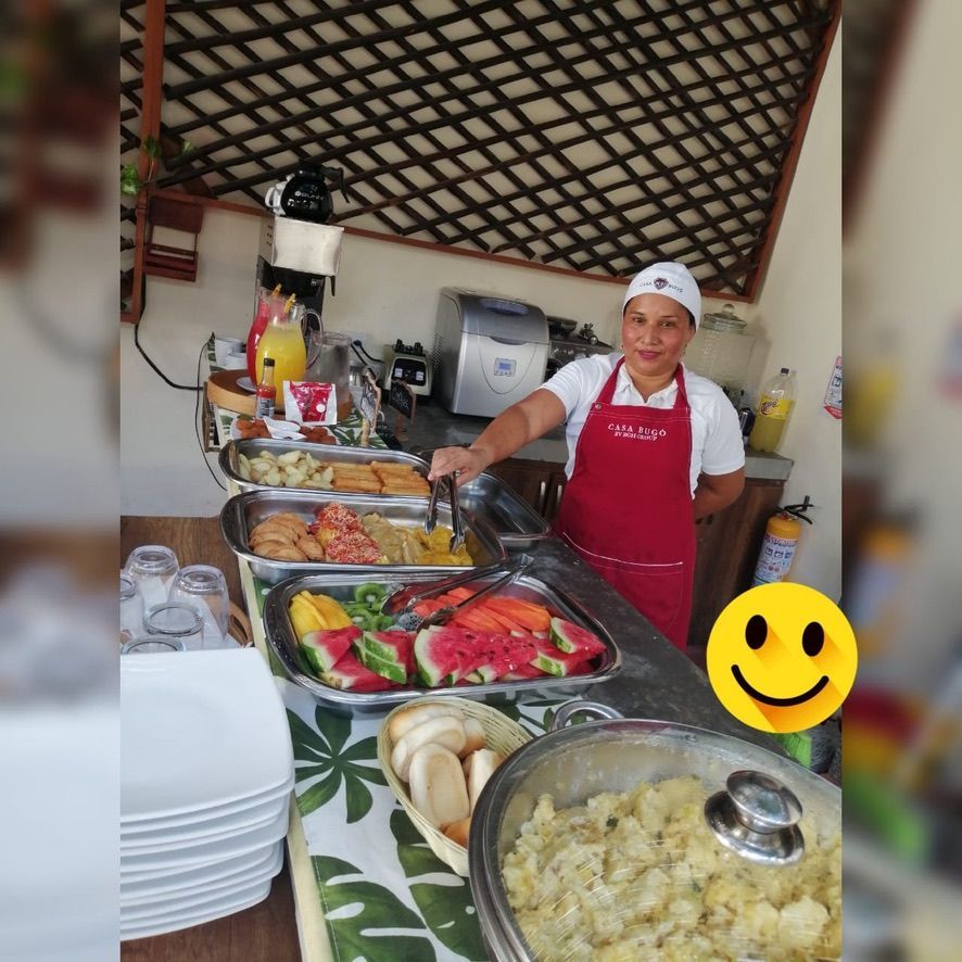 Woman in a red apron serves food at a buffet. Dishes include fruit, potatoes, and other cooked items.
