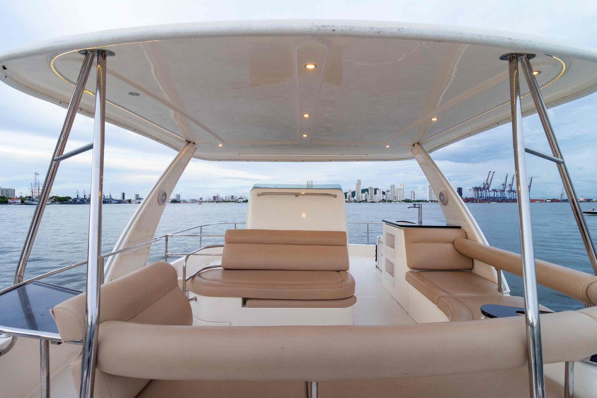 View from inside a boat with beige seating and a canopy, facing the water and a city skyline.