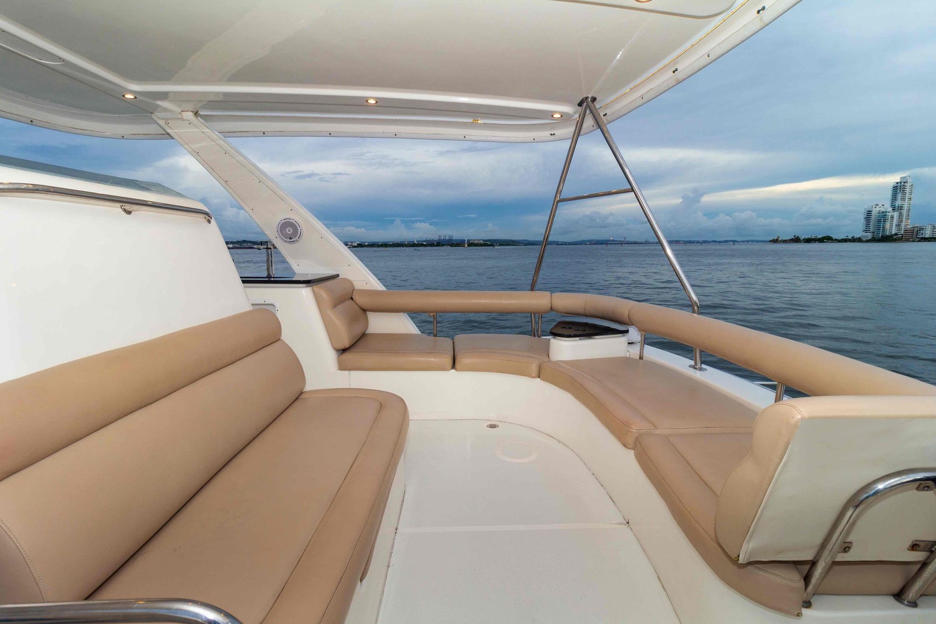 Tan cushioned seating on a white yacht deck, facing the water and cityscape on the horizon under a cloudy sky.