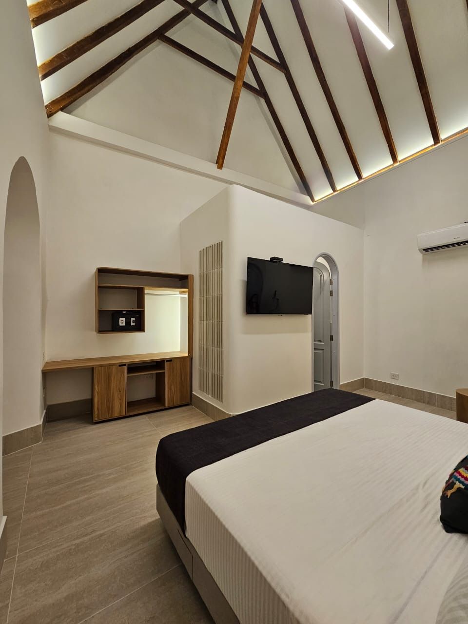 Hotel room with a bed, a desk with a TV, and a vaulted ceiling with exposed beams.