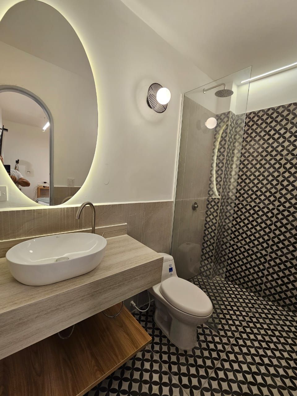 Modern bathroom with an oval-shaped illuminated mirror, vessel sink, and patterned tile flooring. Shower has a waterfall head.