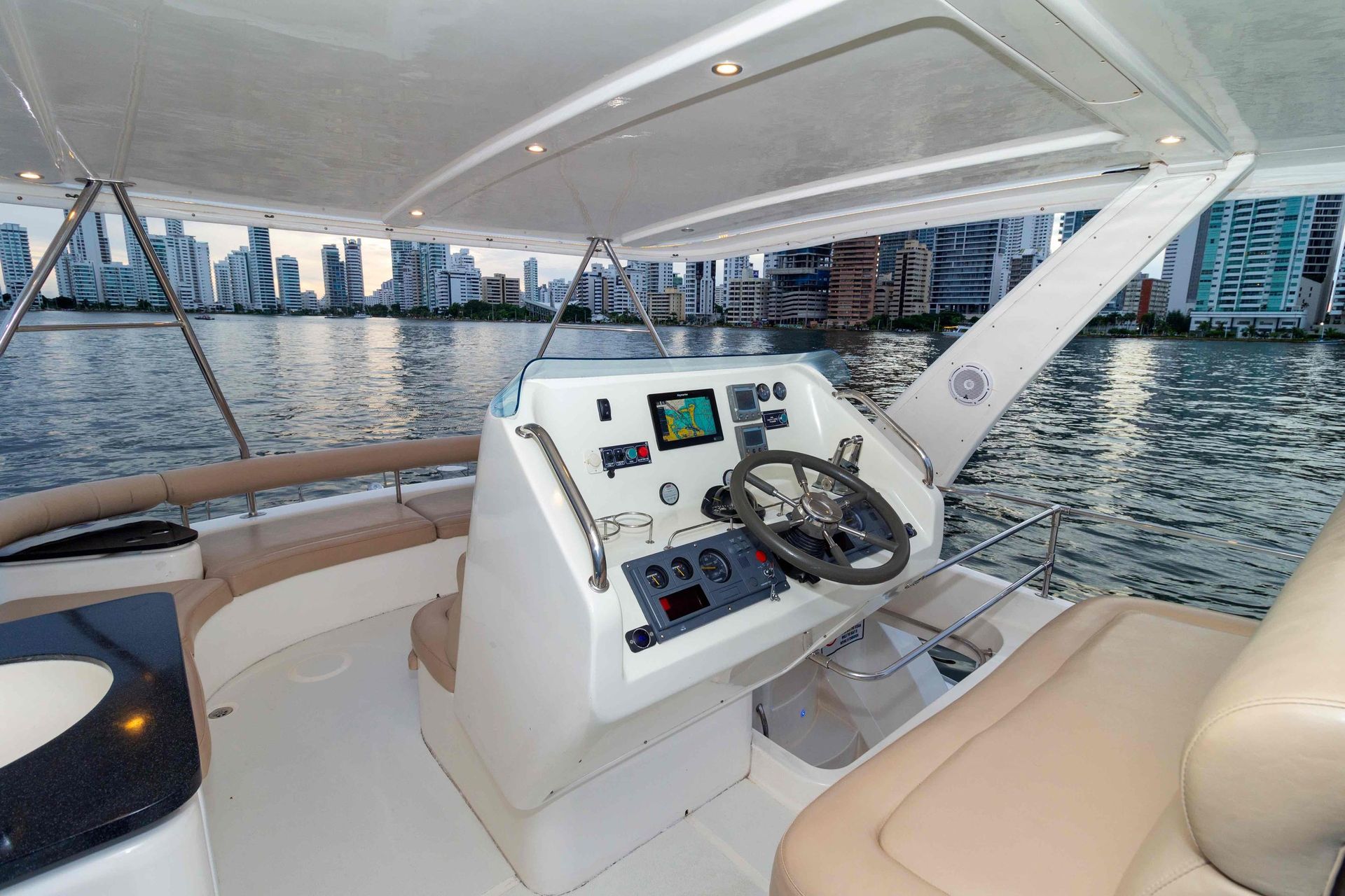 Interior view of a boat's helm with steering wheel and navigation equipment, overlooking a cityscape.