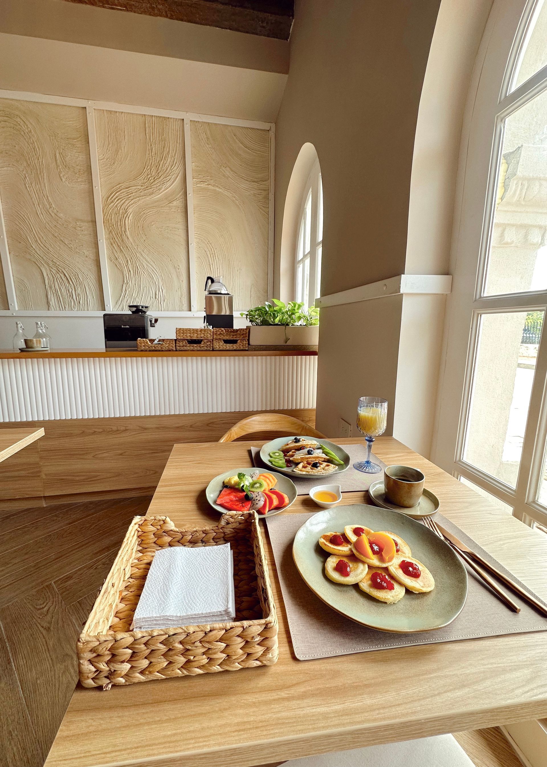 A wooden table set for breakfast near a window with pancakes, fruit, and juice. The room has light walls and a counter in the background.