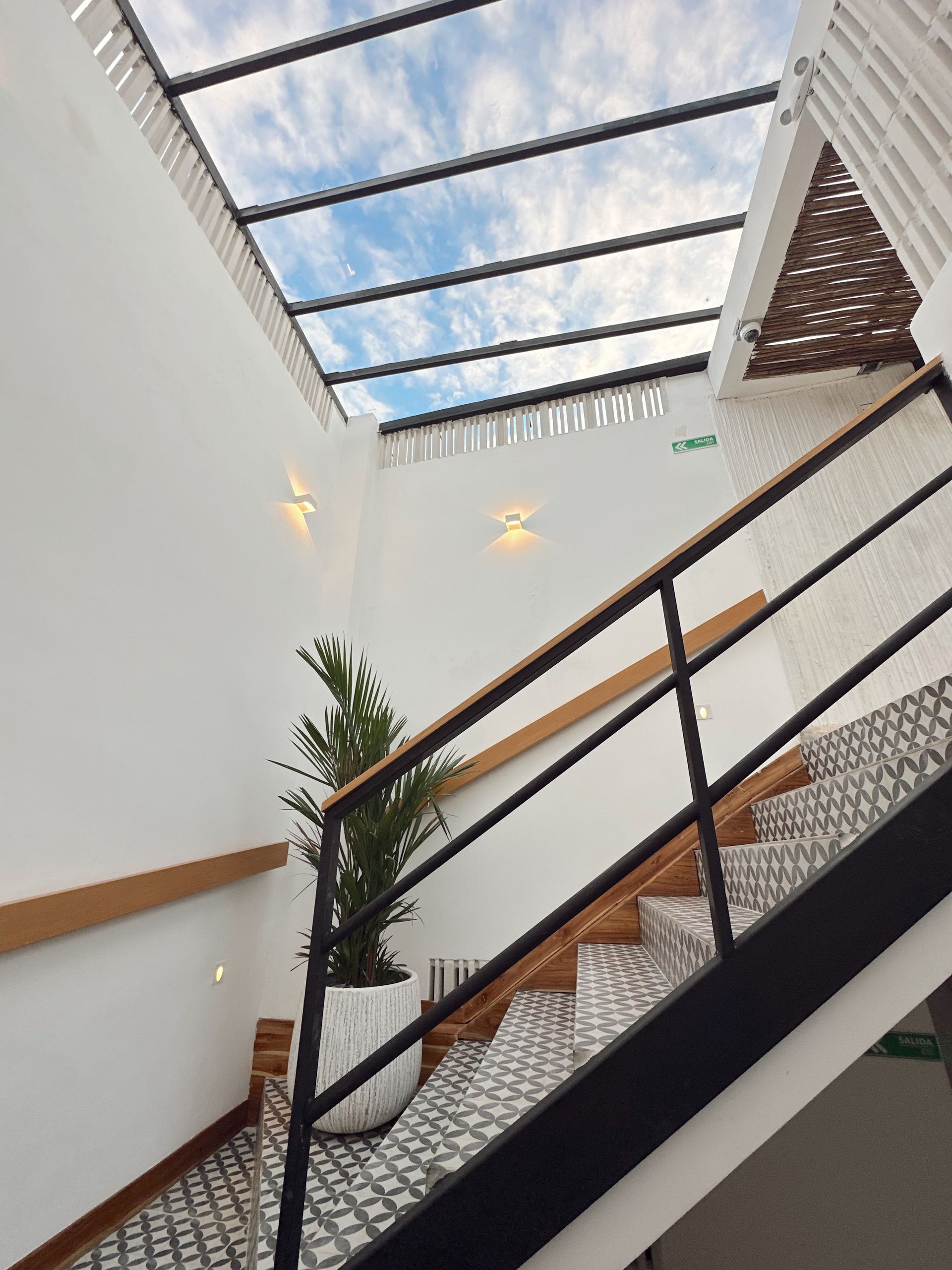 Staircase with black railing and patterned steps leads upward towards a glass ceiling revealing a partly cloudy sky.