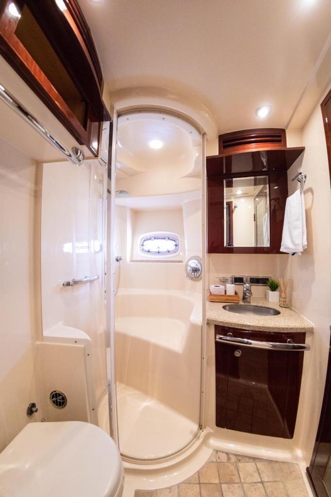 A small, well-lit boat bathroom with a shower, toilet, and sink. Dark wood cabinetry contrasts with the light walls.