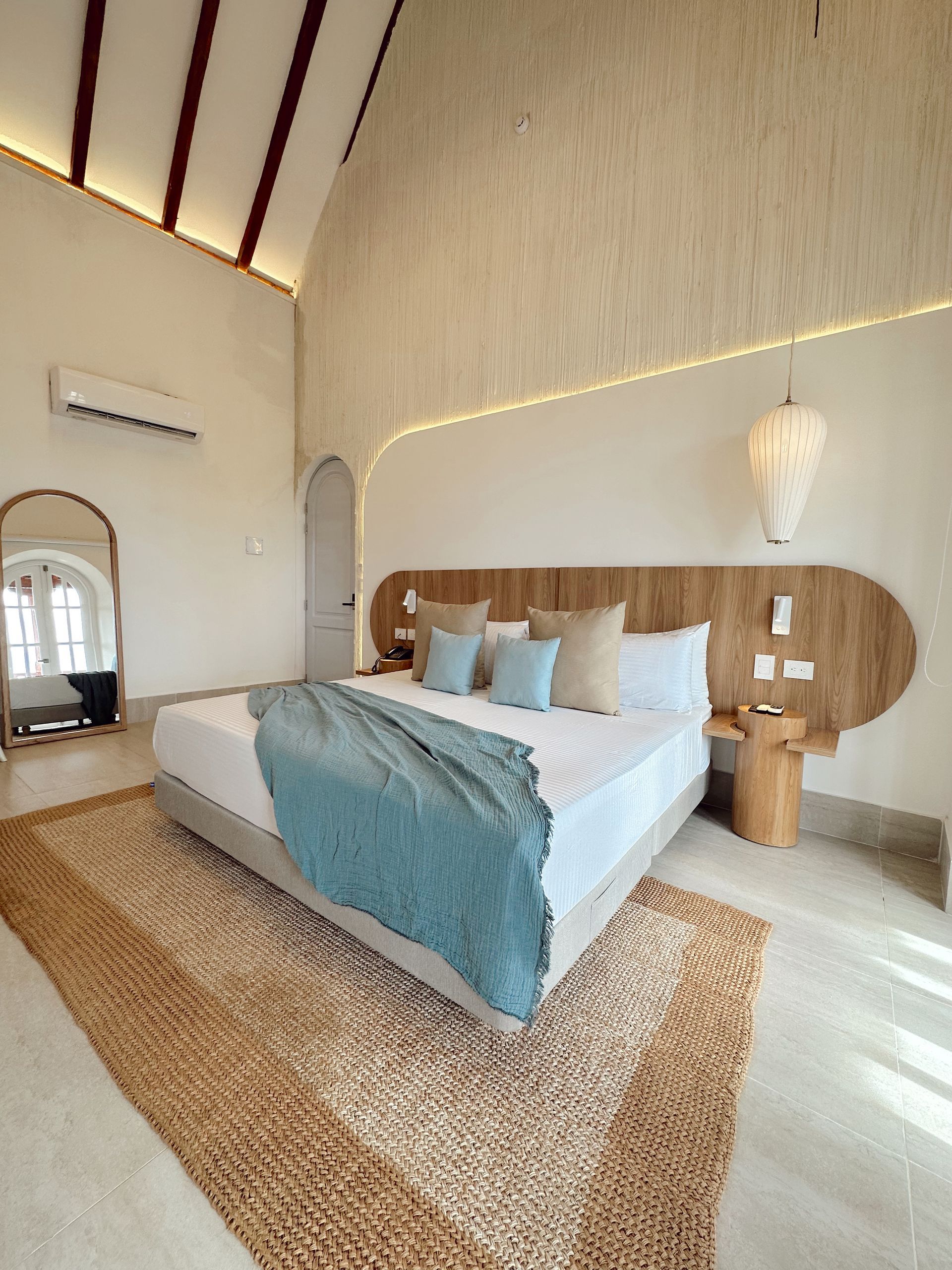 Bedroom with king-size bed, woven rug, light blue throw, wooden headboard, and large arched mirror. Neutral colors and a tropical aesthetic.