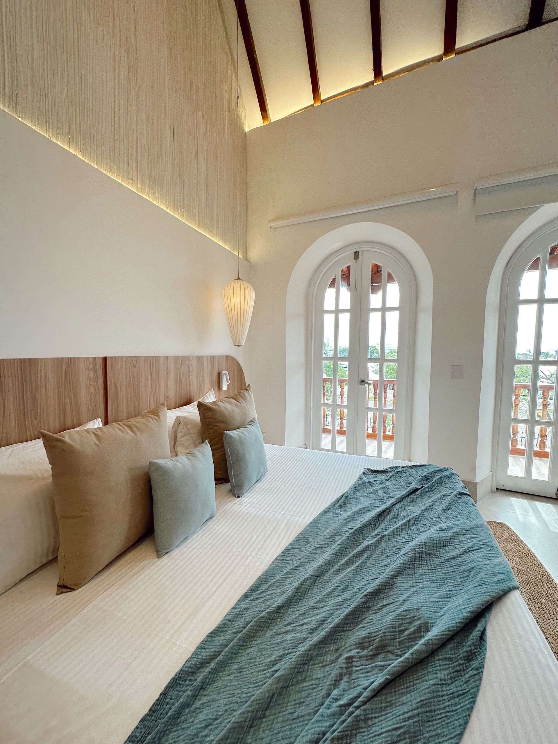 Bright bedroom with a large bed, neutral-colored pillows, a blue blanket, and arched windows. Wooden headboard and high ceiling.