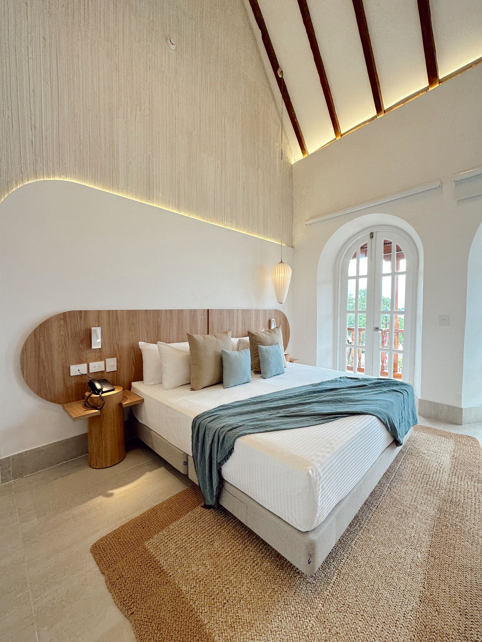 A bright hotel bedroom with a large bed, wooden headboard, and rug. The room has high ceilings and a door to a balcony.