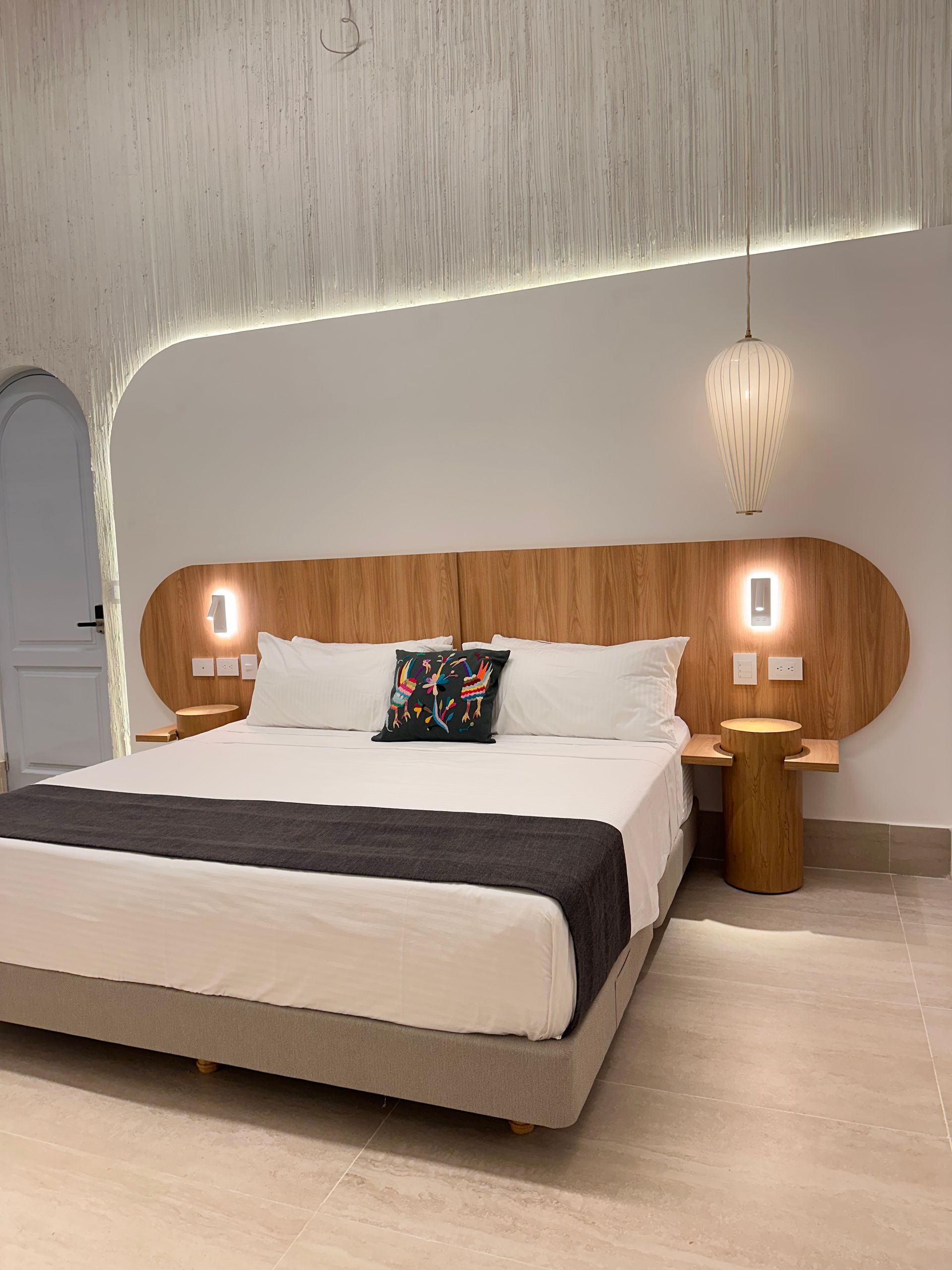 A modern bedroom featuring a large wooden headboard with integrated bedside tables and lamps. A plush bed with white bedding and a decorative pillow is centered.
