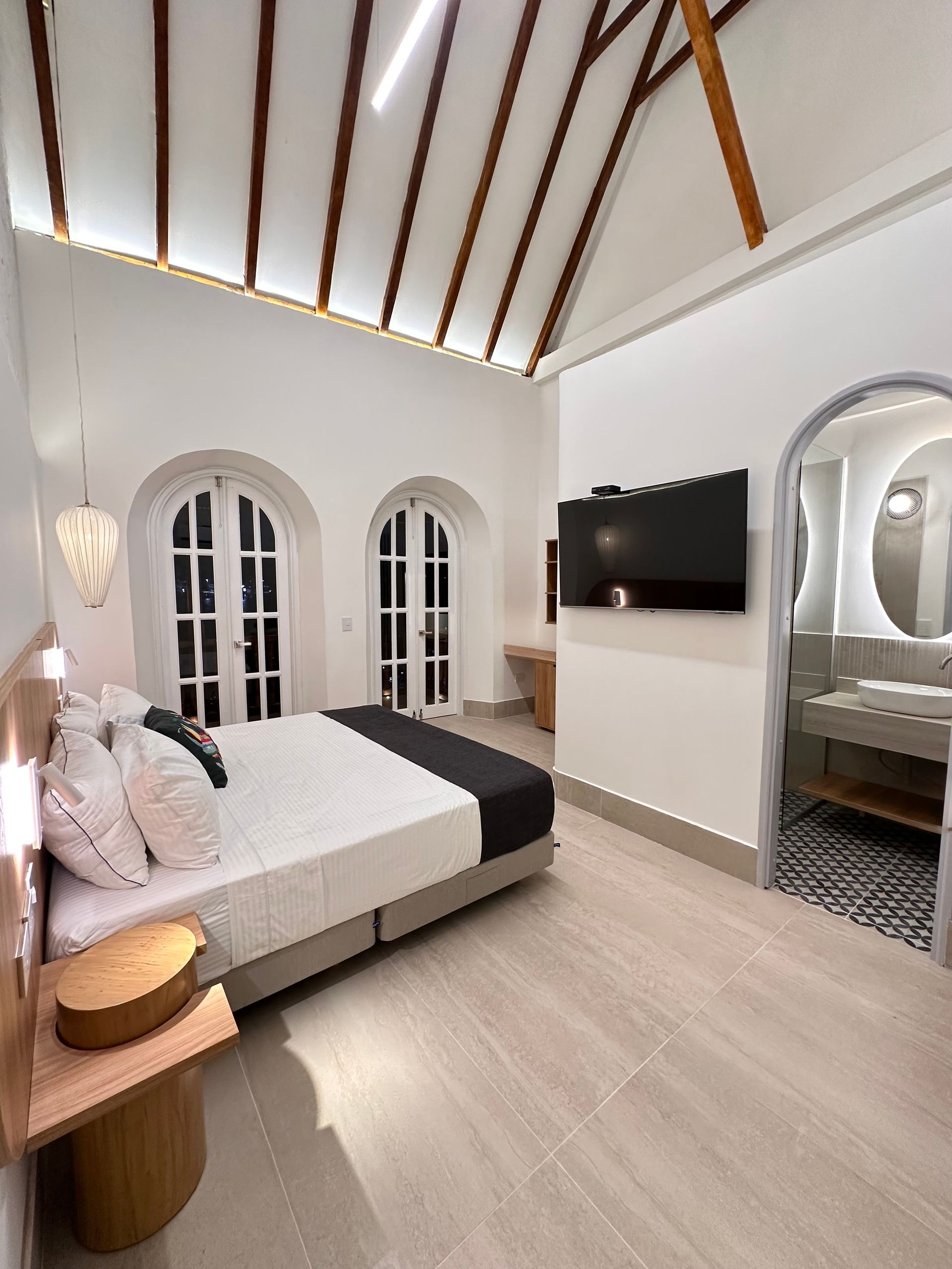 Bedroom with arched windows and door, a bed, and an attached bathroom. White walls, wooden ceiling beams, and a modern design.