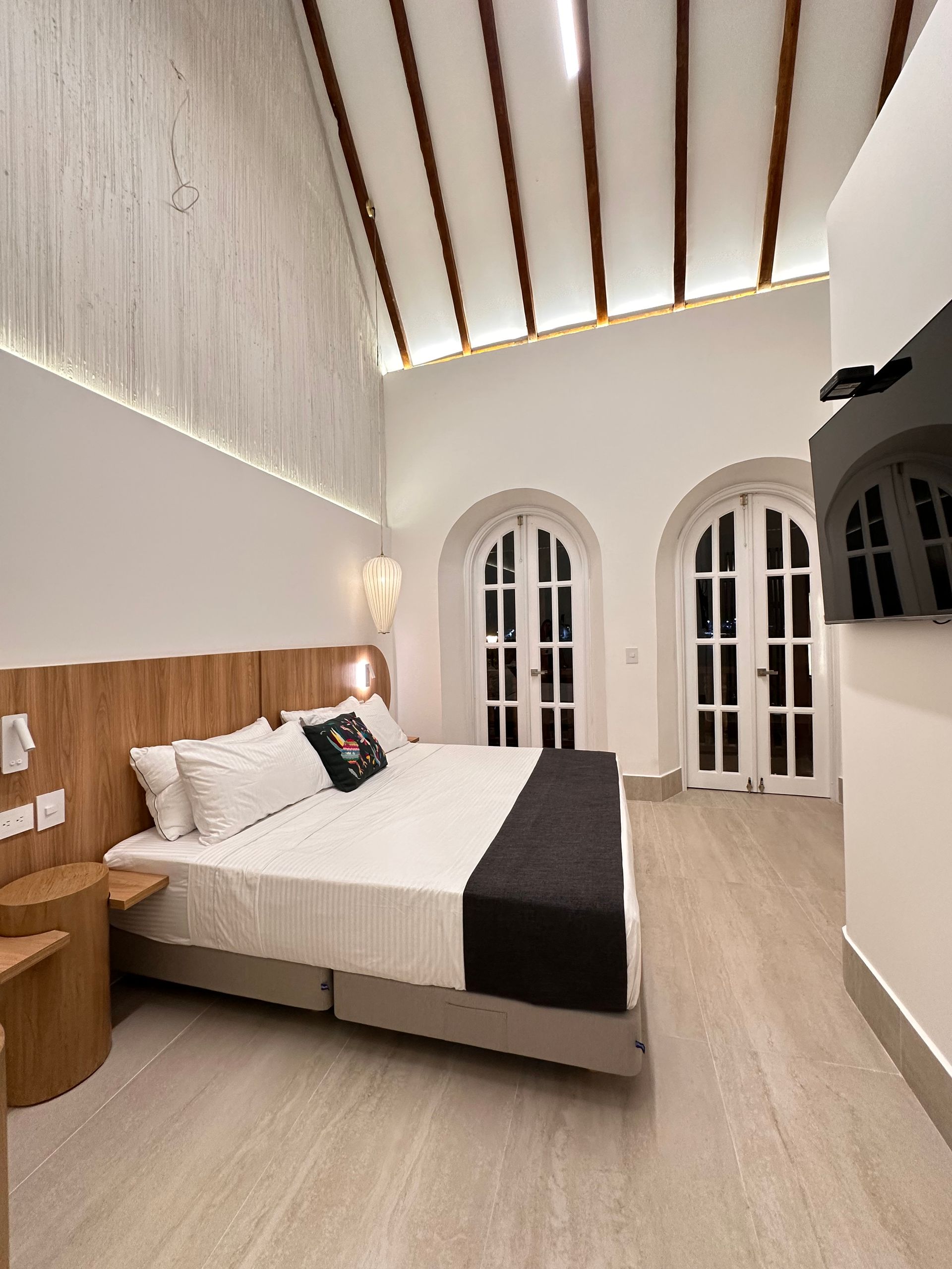A bright hotel bedroom with a large bed, two arched doors, and wooden accents. The room is mostly white with neutral tones.