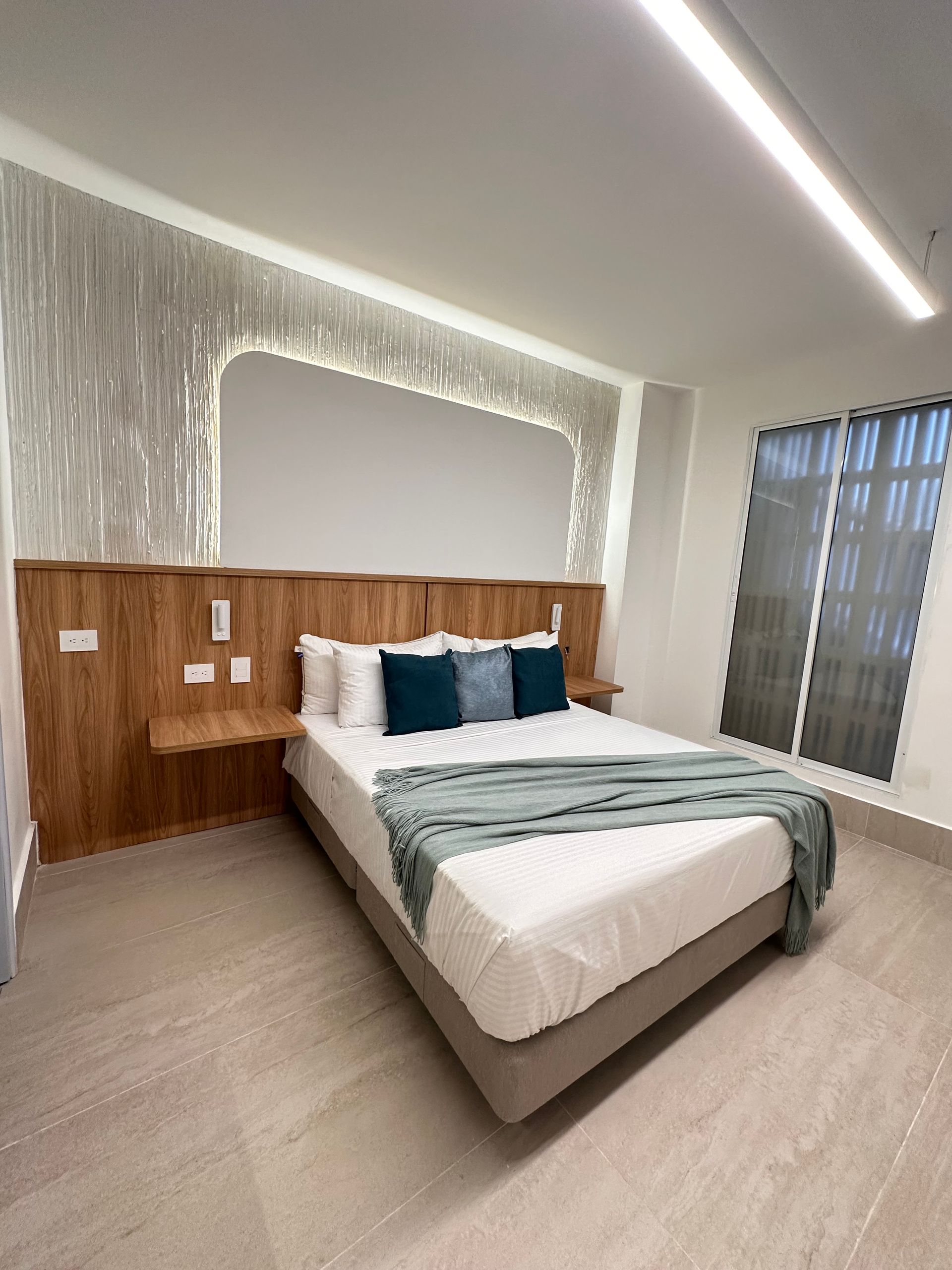 A modern hotel room with a bed centered against a wood panel headboard, teal pillows, and a gray bed skirt.