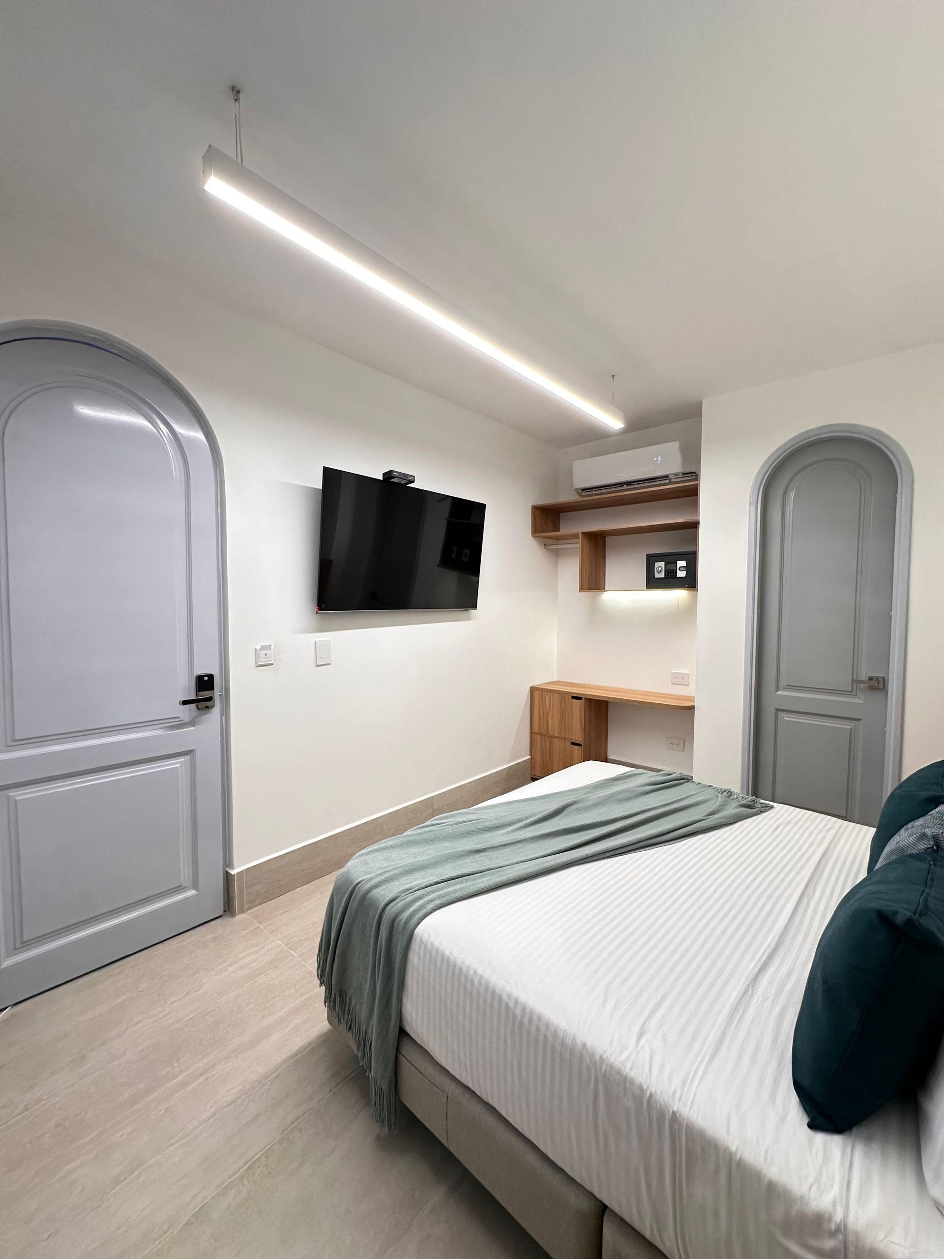 A modern hotel room with a bed, TV, desk, and arched doors. White walls, light wood floors, and a teal blanket are featured.