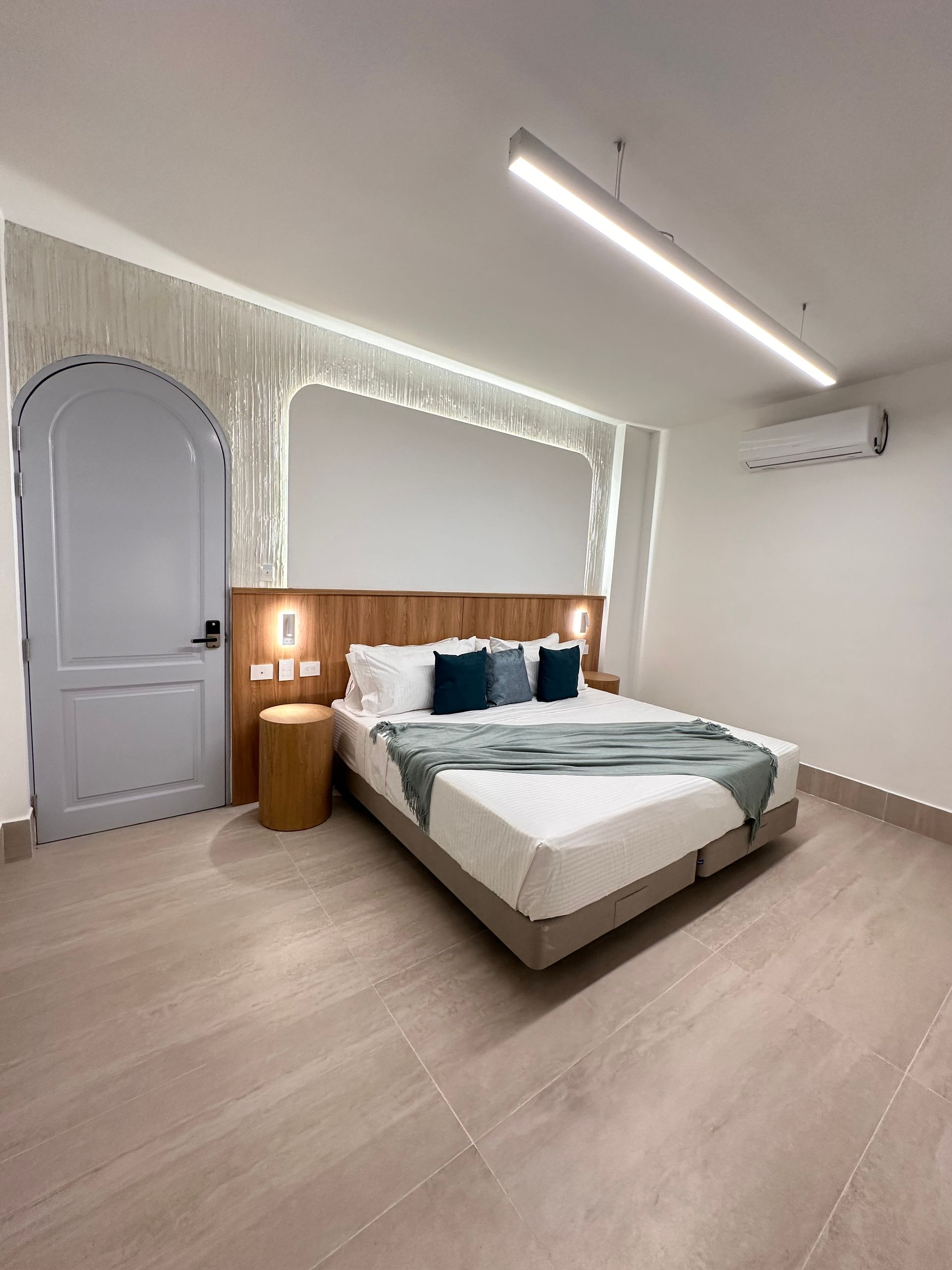Bedroom with a queen-size bed, a side table, and a light-colored door. The room is painted white and has a modern aesthetic.