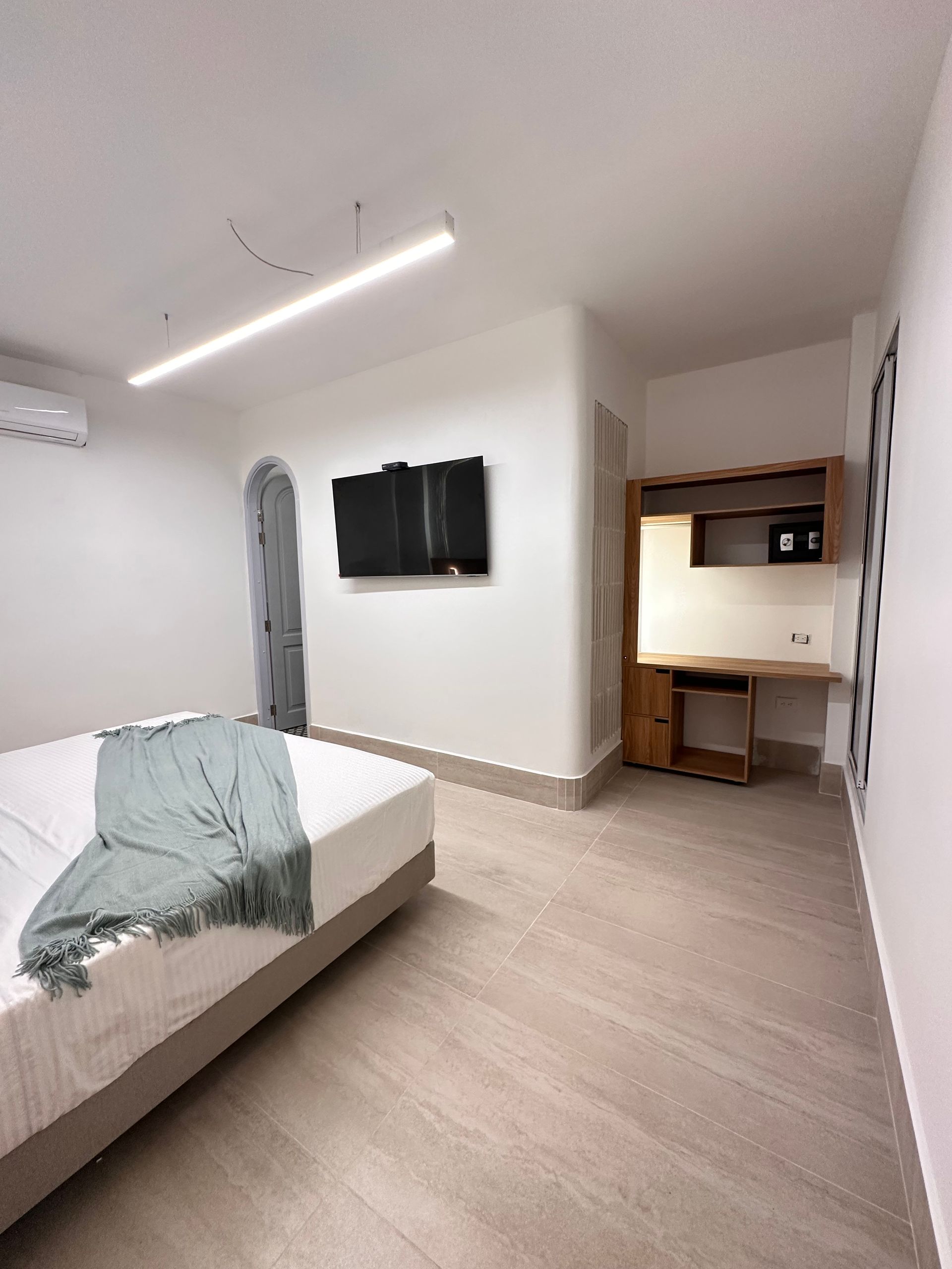 Bright white bedroom with a bed, TV, desk, and archway. Soft blue blanket, and light-colored wooden furniture.
