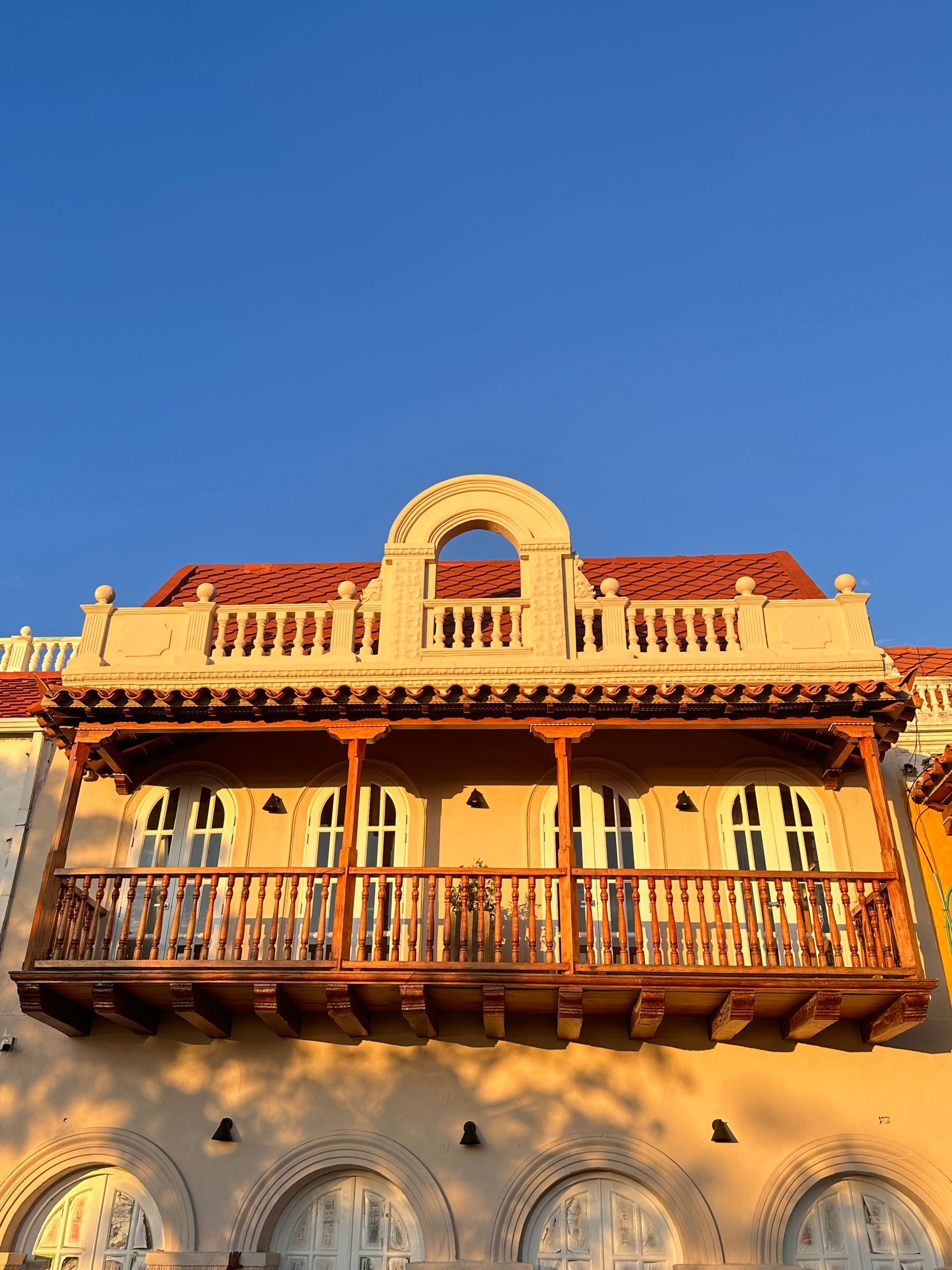 Balcony of a building in Cartagena, Colombia. The wooden balcony is brown with a white stucco facade and a blue sky above.