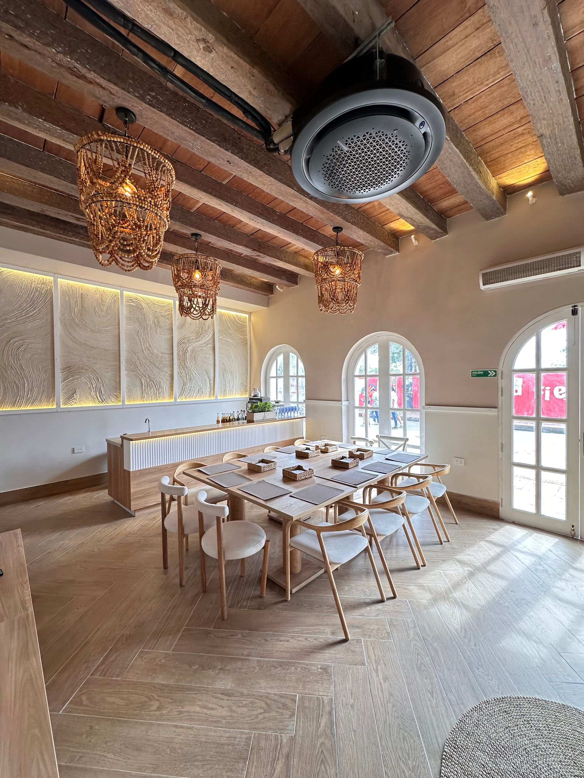 Bright dining room with a long table set for a meal, adorned with woven lamps and a high ceiling with wooden beams.
