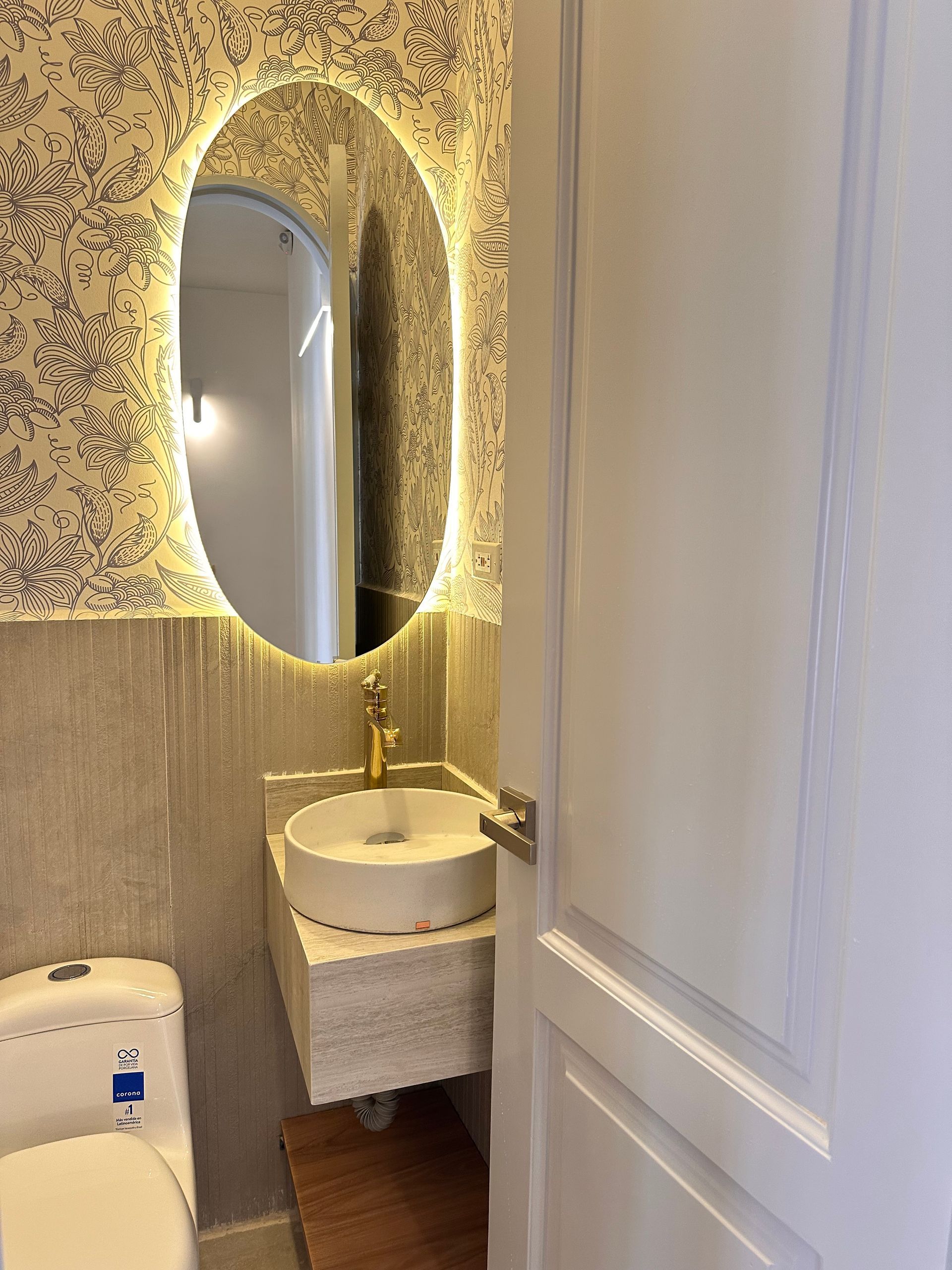 Small bathroom with oval mirror, lit up around the edges, above a white sink and golden faucet. A white toilet is on the left.