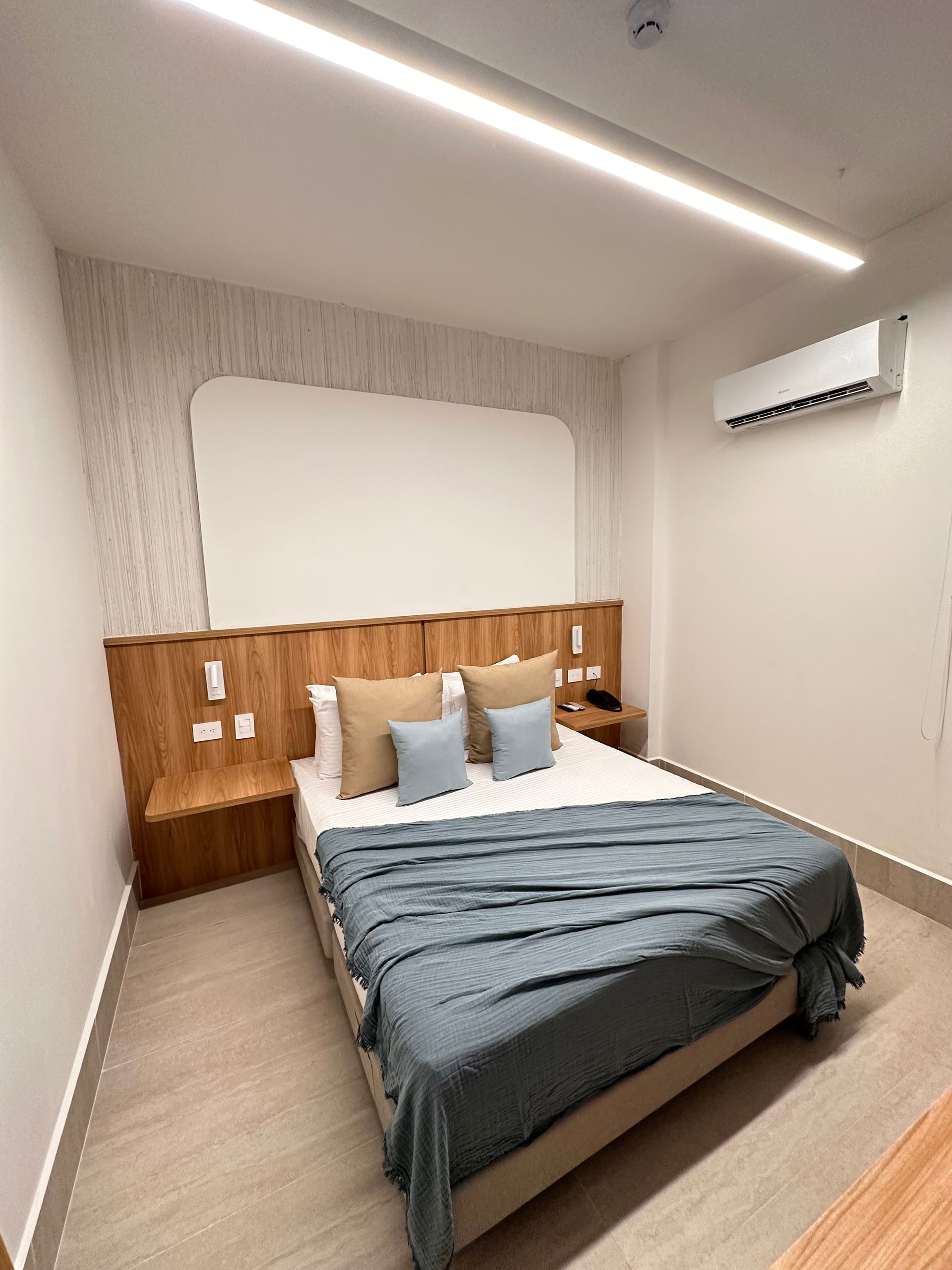 Hotel bedroom with a wooden headboard, bed with blue blanket, light-colored walls, and air conditioning.